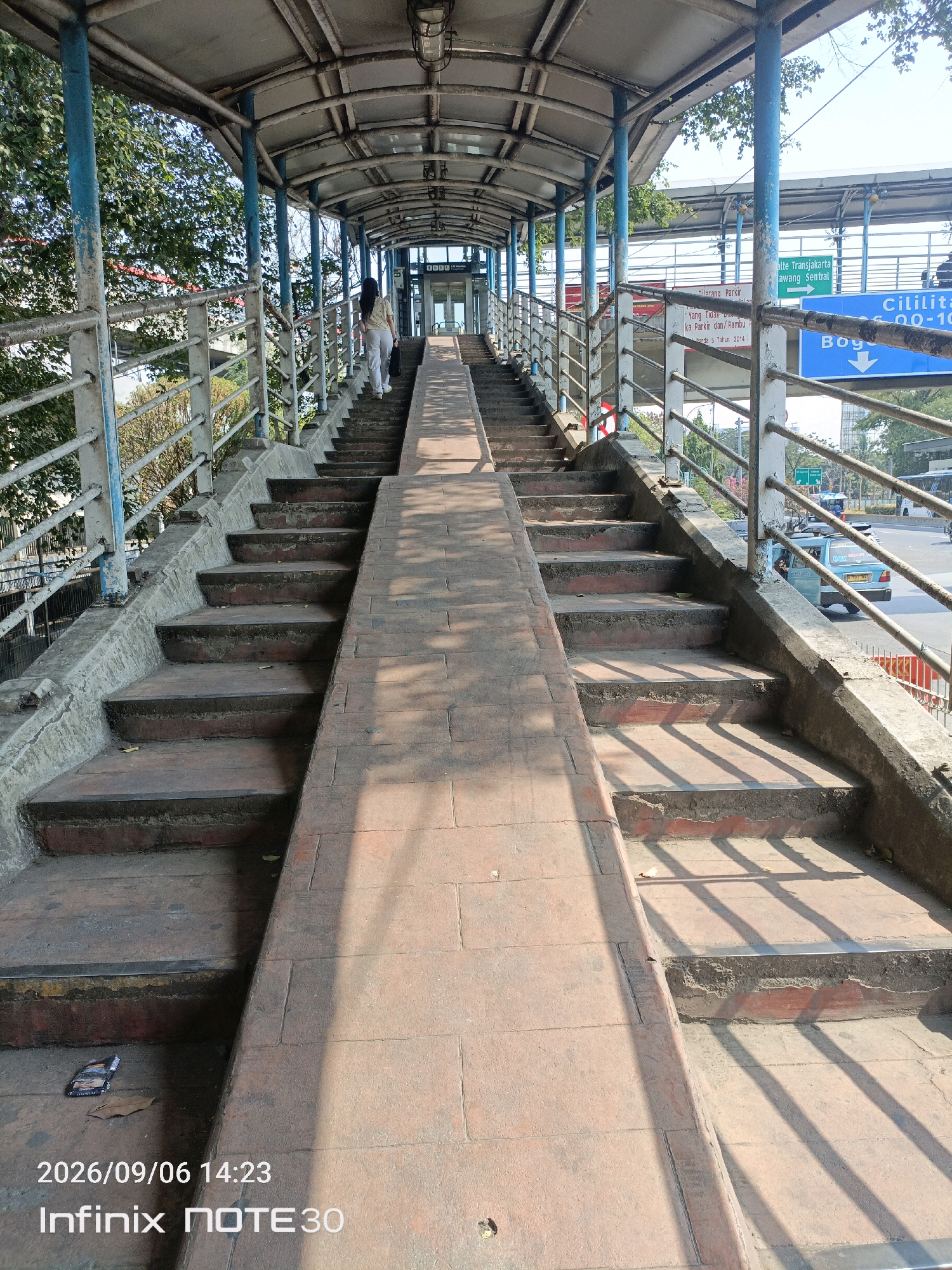 The image shows an elevated pedestrian walkway in East Jakarta, Indonesia, likely a pedestrian bridge over a road. The walkway is covered by a metal roof supported by blue pillars. On the left side, a woman in a light-colored shirt and pants is walking up a flight of stairs. The stairs are made of concrete with red brick risers. To the right of the stairs, there is a ramp made of brick tiles.
Overhead signs are visible, one with green text reading "Ilte Transjakarta Awang Sentral" and another with blue text reading "Cililitan 00-10". The environment appears to be urban, with trees visible on the left and traffic on the road below on the right. The walkway shows signs of wear and tear, with some rust on the railings and chipped concrete. The bottom of the image displays a timestamp "2026/09/06 14:23" and phone model "Infinix NOTE30".