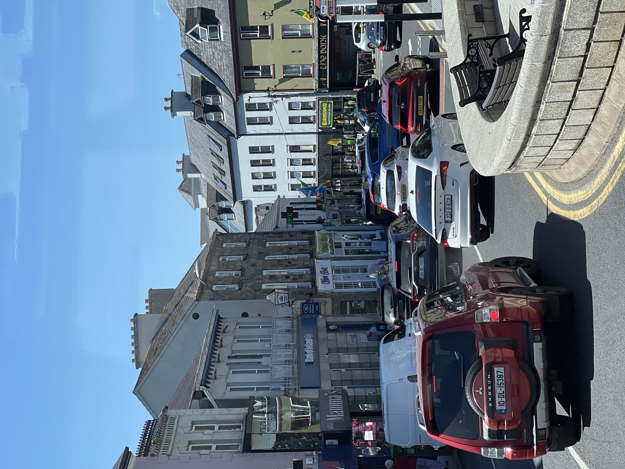 The image shows a street scene in Donegal, Ireland, on a sunny day. Several cars are stopped in traffic on a paved road with yellow lane markings. Buildings line both sides of the street, featuring a mix of architectural styles and colors. Some buildings have signage indicating businesses such as "Bank of Ireland," "Mamma's," "Little," "Eurosaver," and "The Bank." People are visible on the sidewalks and in some of the cars. The sky is clear and blue. A stone wall with a bench is in the foreground.