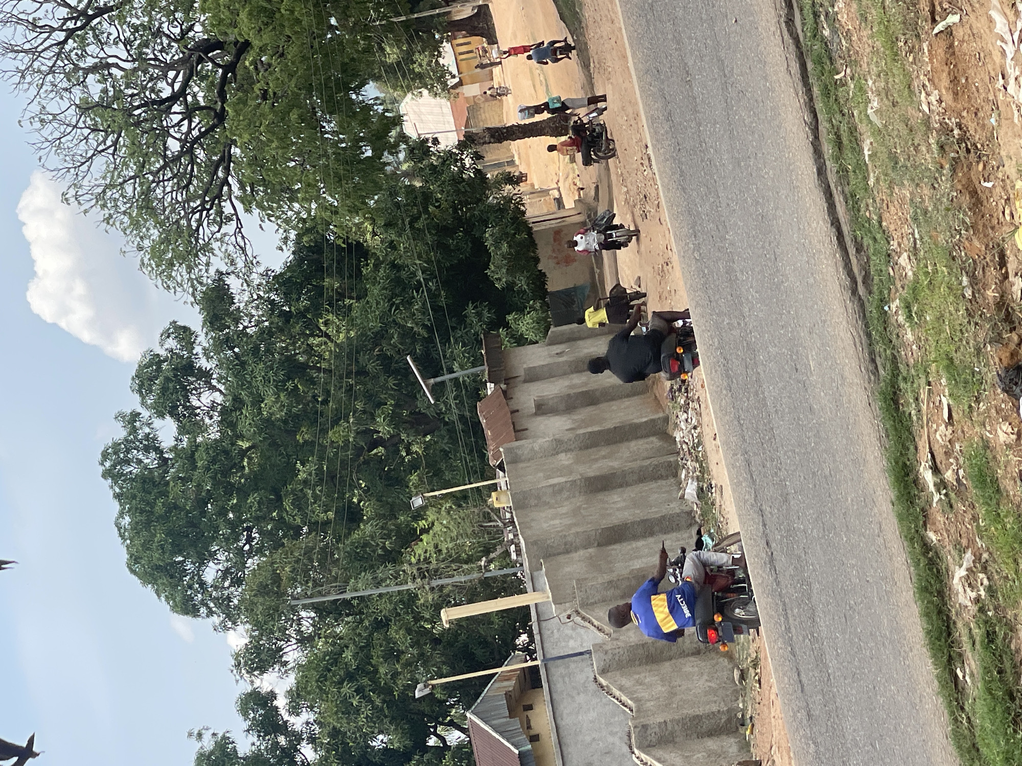 This image captures a vibrant outdoor street scene in Mararaba, Nigeria, during daylight hours. The sky is clear with a prominent white cloud, and abundant green trees dominate the background and left side of the frame.
A paved road traverses the middle ground, bordered on one side by an unpaved embankment strewn with grass and litter. On the opposite side, a series of unfinished concrete stairs ascends alongside the road. Several people, mostly men, are visible throughout the scene, primarily on or near motorcycles. In the foreground, a man wearing a blue and yellow shirt with "DIRECTV" printed on the back is seated on a motorcycle next to the stairs. Further along, another man in a black shirt is also on a motorcycle. Other individuals are seen walking or standing in the background near simple buildings, some with corrugated iron roofs. Utility poles with overhead wires are also present, indicating basic infrastructure. The scene depicts typical daily activity in a semi-urban or rural setting.