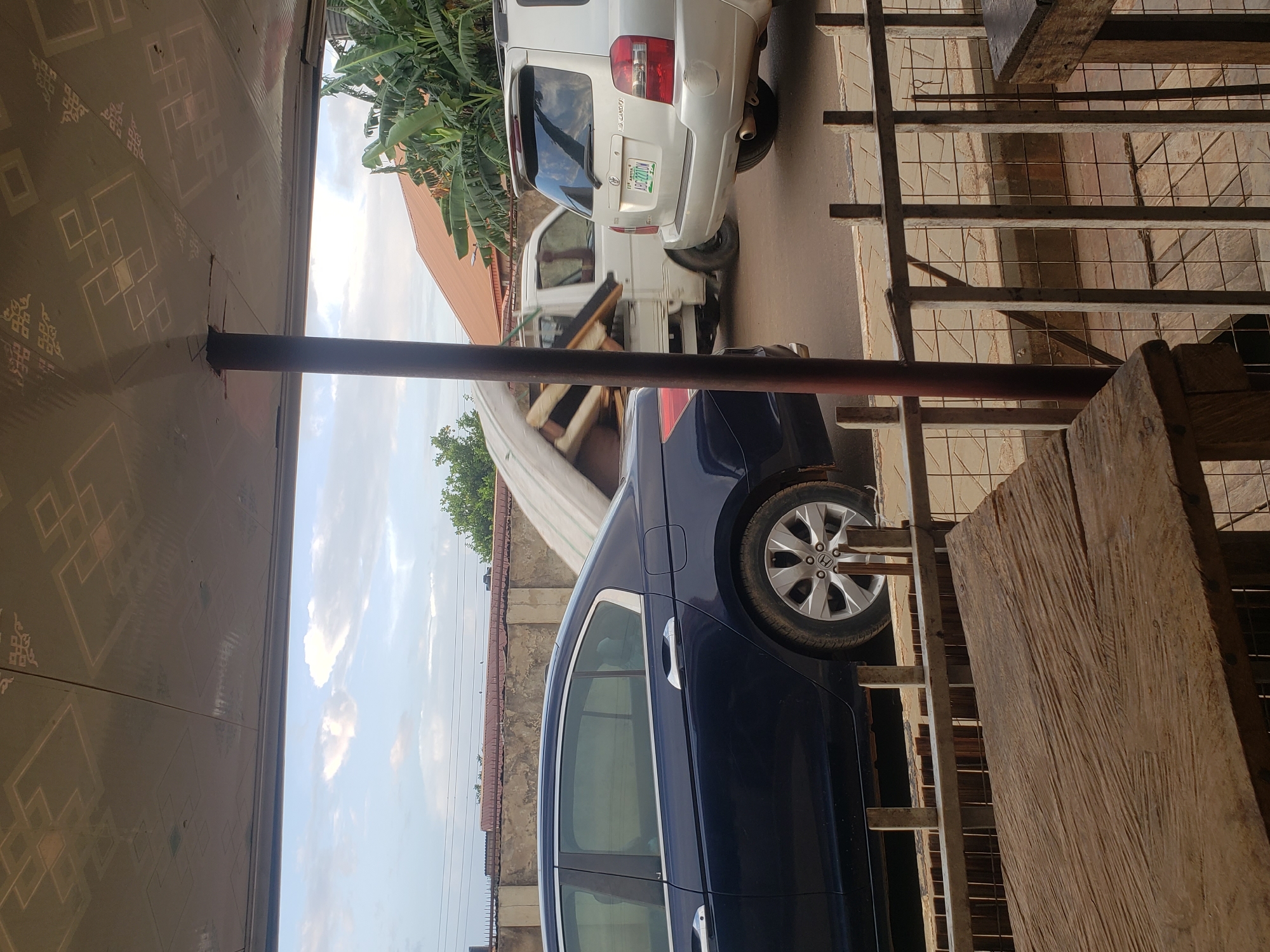 The image captures a street scene in Narayi, Nigeria, from a slightly elevated, covered vantage point, possibly a veranda. In the foreground, a rough-hewn wooden table and a metal railing with wire mesh backing suggest an outdoor seating area. Beyond the railing, a dark blue sedan is parked on the street. Further down the road, a white truck is loaded with what appears to be disassembled furniture or mattresses, partially covered by a white tarp, and it is in motion. To the right of the truck, a silver Toyota Highlander SUV is also parked on the street. The background features a textured concrete wall, a terracotta-tiled roof, and lush green banana trees, indicating a tropical or subtropical environment. The sky above is partly cloudy. The overall impression is of a quiet street with vehicles and some casual outdoor furniture.