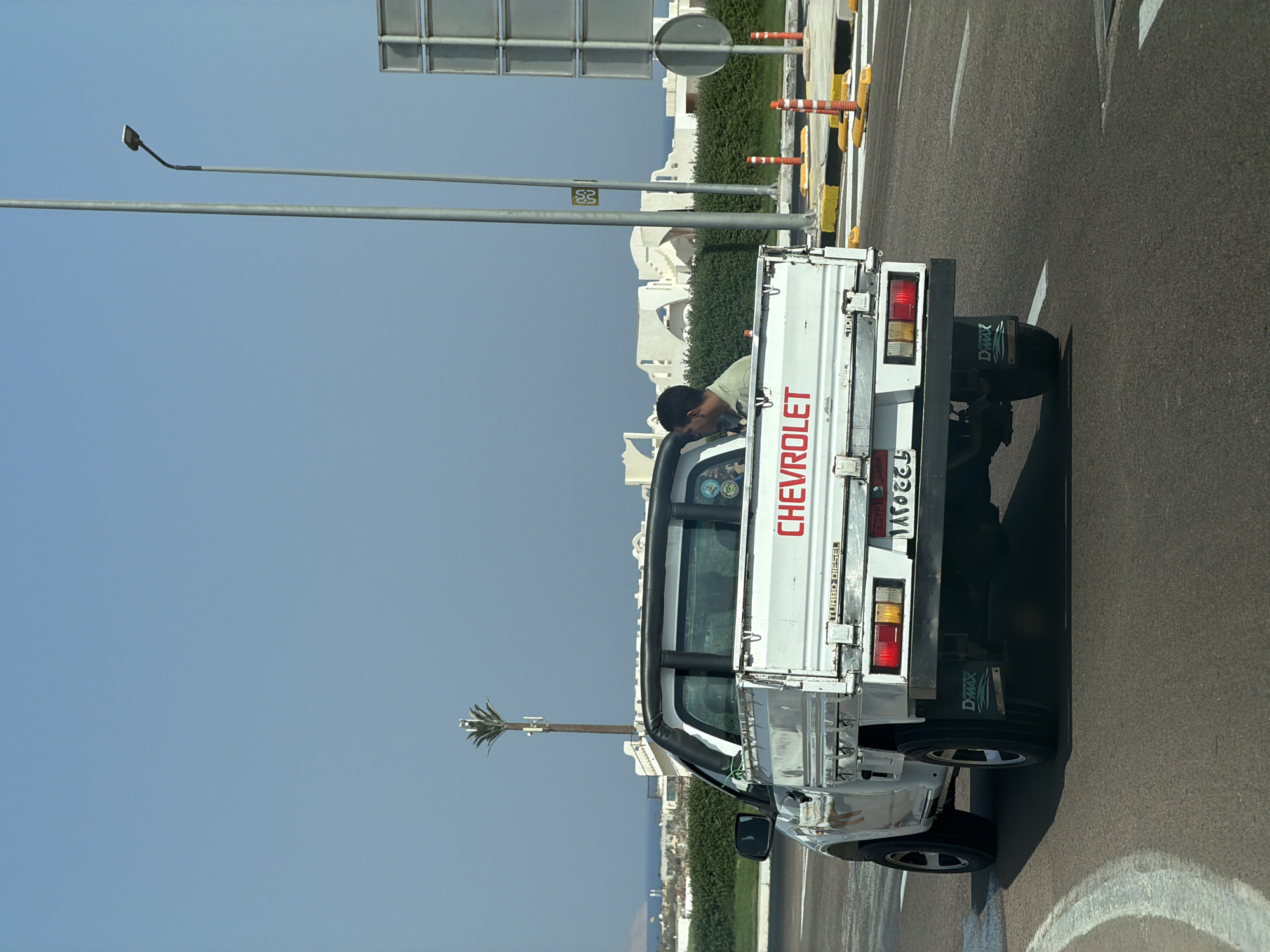 A white Chevrolet pickup truck is driving on a paved road under a clear blue sky in Sharm El Sheikh, Egypt. The truck has a black roll bar and a black trim around the top of the cab. A man wearing a light green shirt is visible in the driver's seat. The truck has a license plate with Arabic characters. In the background, there are white buildings with arched architecture, green bushes, a palm tree with cell towers, and streetlights. The road has yellow and white markings, and there are orange traffic cones and yellow speed bumps on the side.