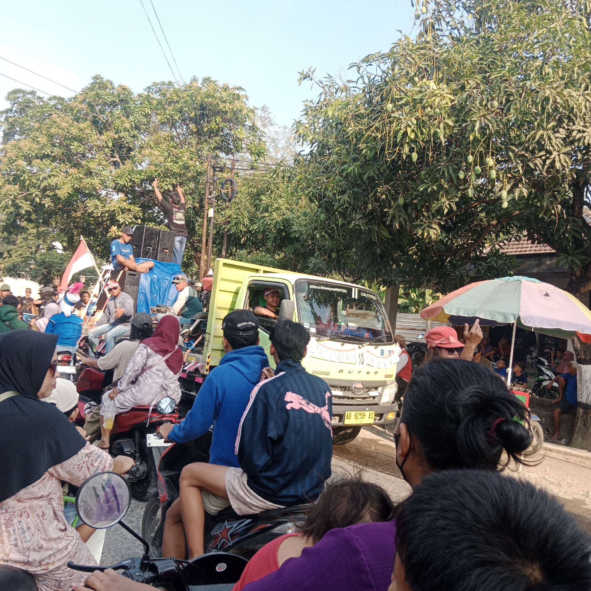 The image depicts a street scene in Tegalrejo, Indonesia, likely during a procession or gathering. A bright yellow truck, possibly a flatbed, serves as a central element, adorned with decorations and carrying several people and audio equipment, including large speakers. A flag of Indonesia is visible, adding to the festive or patriotic atmosphere. Numerous individuals are present, many on motorcycles, some standing on the truck, and others in the foreground, observing the event. The environment is characterized by trees with dense foliage, a clear blue sky, and a paved road. The presence of an umbrella suggests a sunny day. People are dressed in casual attire, with some wearing headscarves. The overall impression is one of a lively community event or celebration.