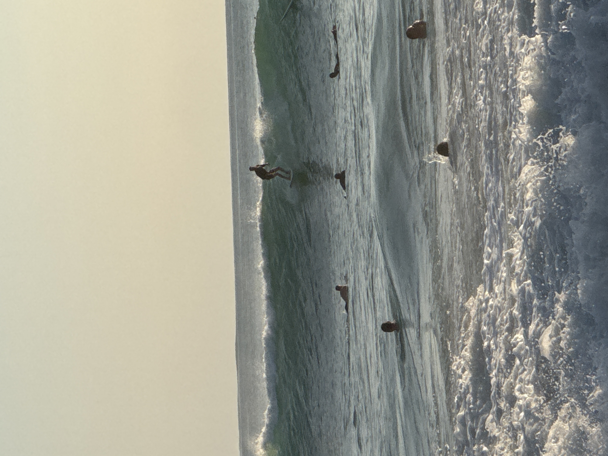 The image depicts surfers on a wave in Kuta Selatan, Indonesia. A surfer is seen riding the wave, while others are in the water nearby. The wave is breaking with white foam. The sky is overcast.