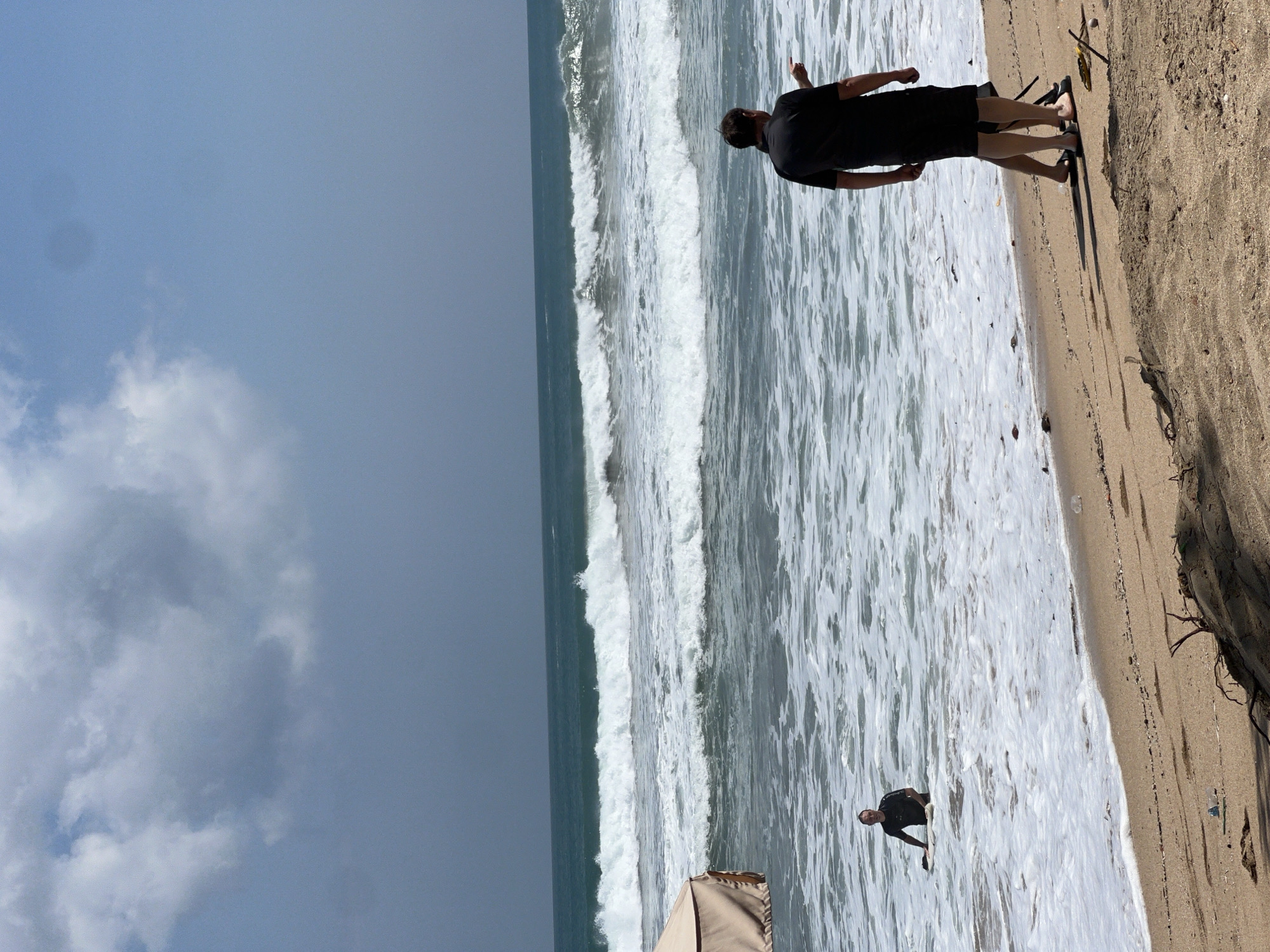 The image depicts a scene at the beach in Kuta, Indonesia. In the water, a man in a black wetsuit is sitting on a surfboard. Further out, large waves are breaking. On the sandy shore, two men are standing, with one pointing towards the ocean. A tan-colored beach umbrella is visible to the left. The sky is partly cloudy.