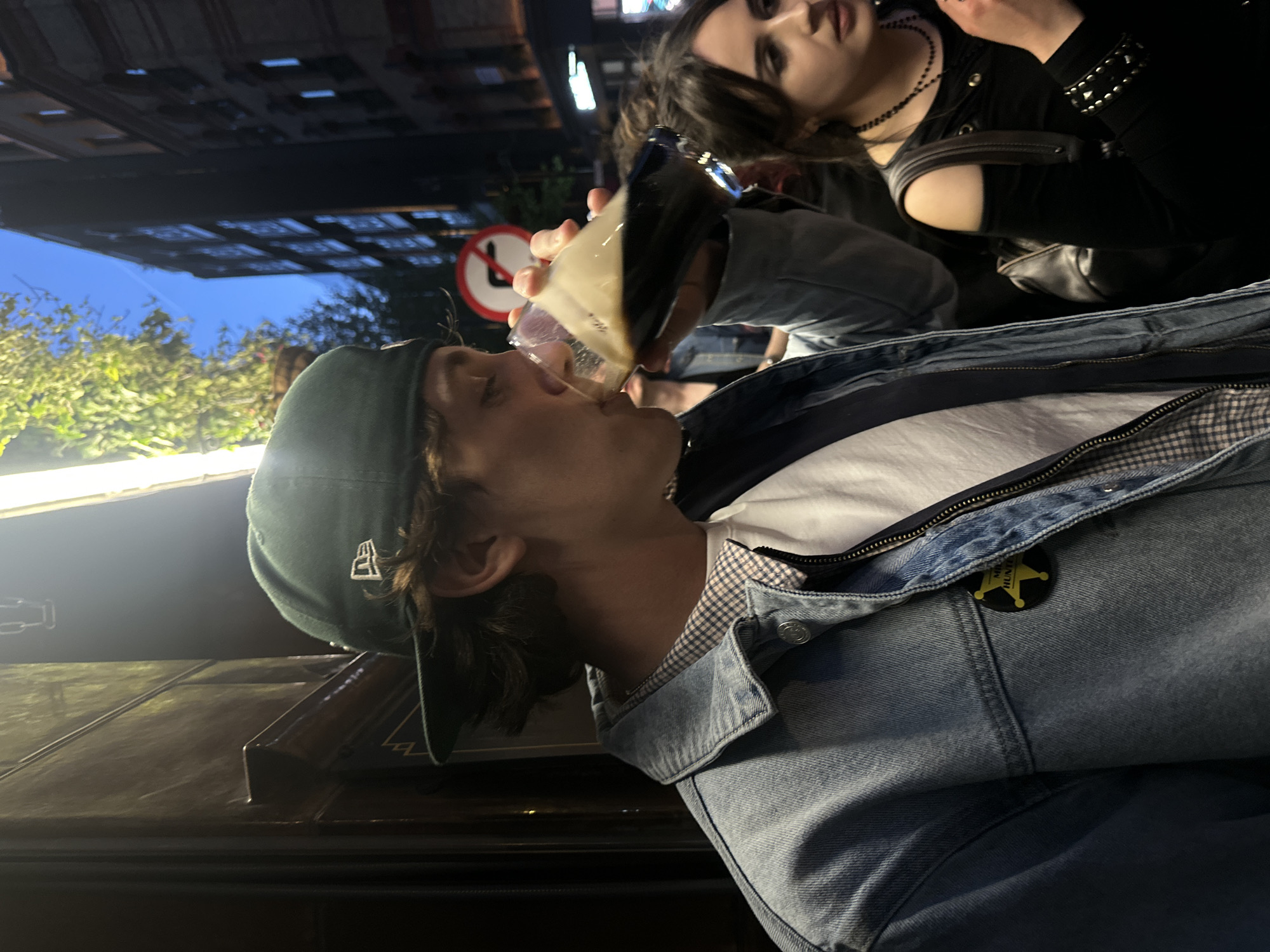 A low-angle, poorly-lit shot depicts a fair-skinned young man drinking from a glass or mug, with a fair-skinned young woman visible in the background. The man is wearing a dark-green baseball cap, a light-blue denim jacket over a white t-shirt, and a gray and white checkered collared shirt underneath. A yellow star-shaped pin is affixed to the denim jacket. The woman has dark hair and is wearing a black top and choker necklace. Both individuals appear to be positioned outdoors in an urban environment. A street sign is visible in the background. Based on the orientation of the street sign relative to the subject, the image has been rotated clockwise.