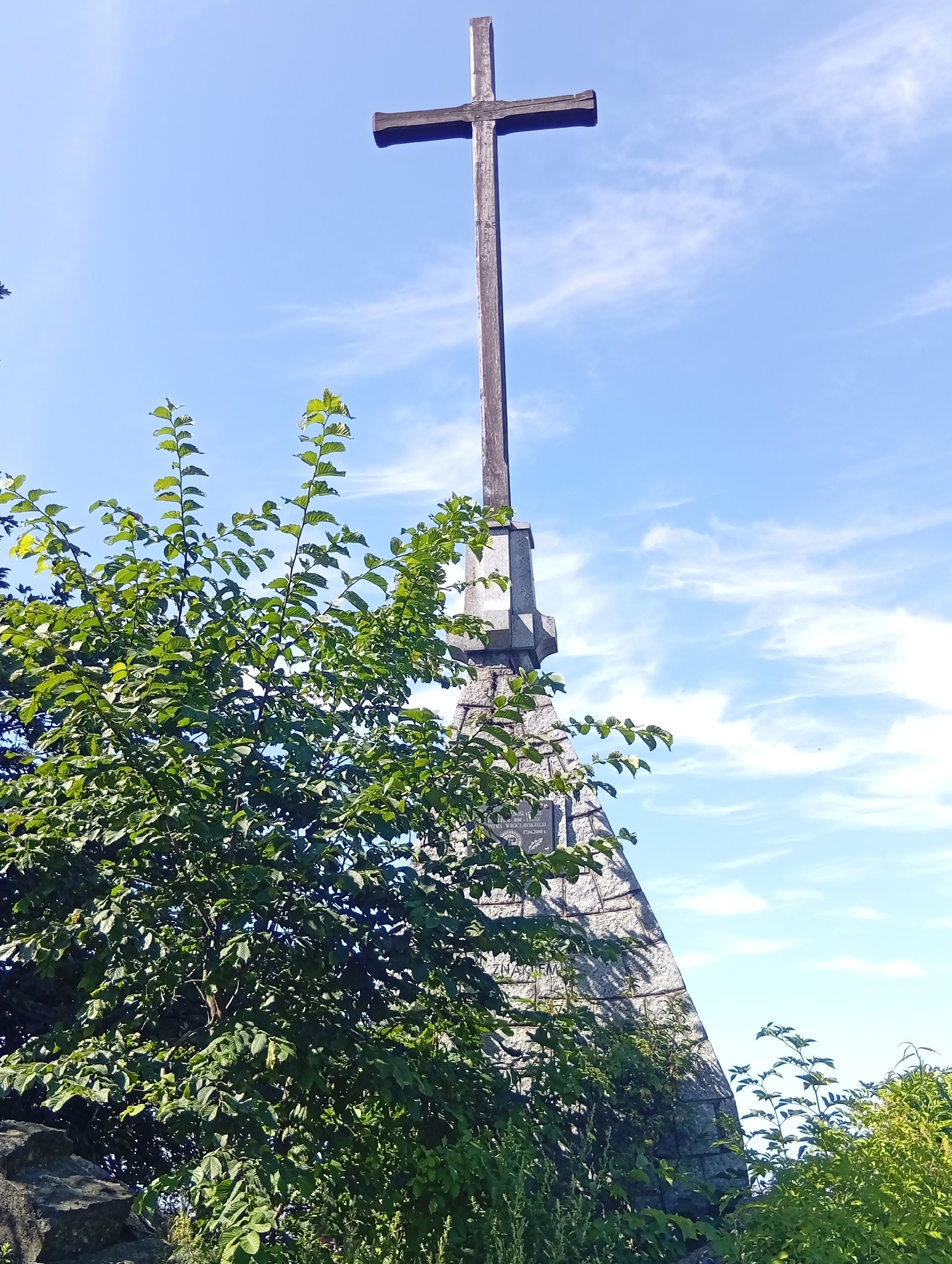 A wooden cross stands atop a stone structure against a bright blue sky with scattered white clouds. The cross is dark brown and appears weathered, with a vertical post and a shorter horizontal beam. The stone structure is triangular in shape, made of rough, light-colored stones and appears to be part of a monument. Lush green foliage, likely trees and bushes, obscures the lower portion of the stone structure and extends from the left side of the frame, reaching towards the cross. The top of a portion of the cross is visible in the top center of the image. The sky is a clear blue with thin, wispy clouds.