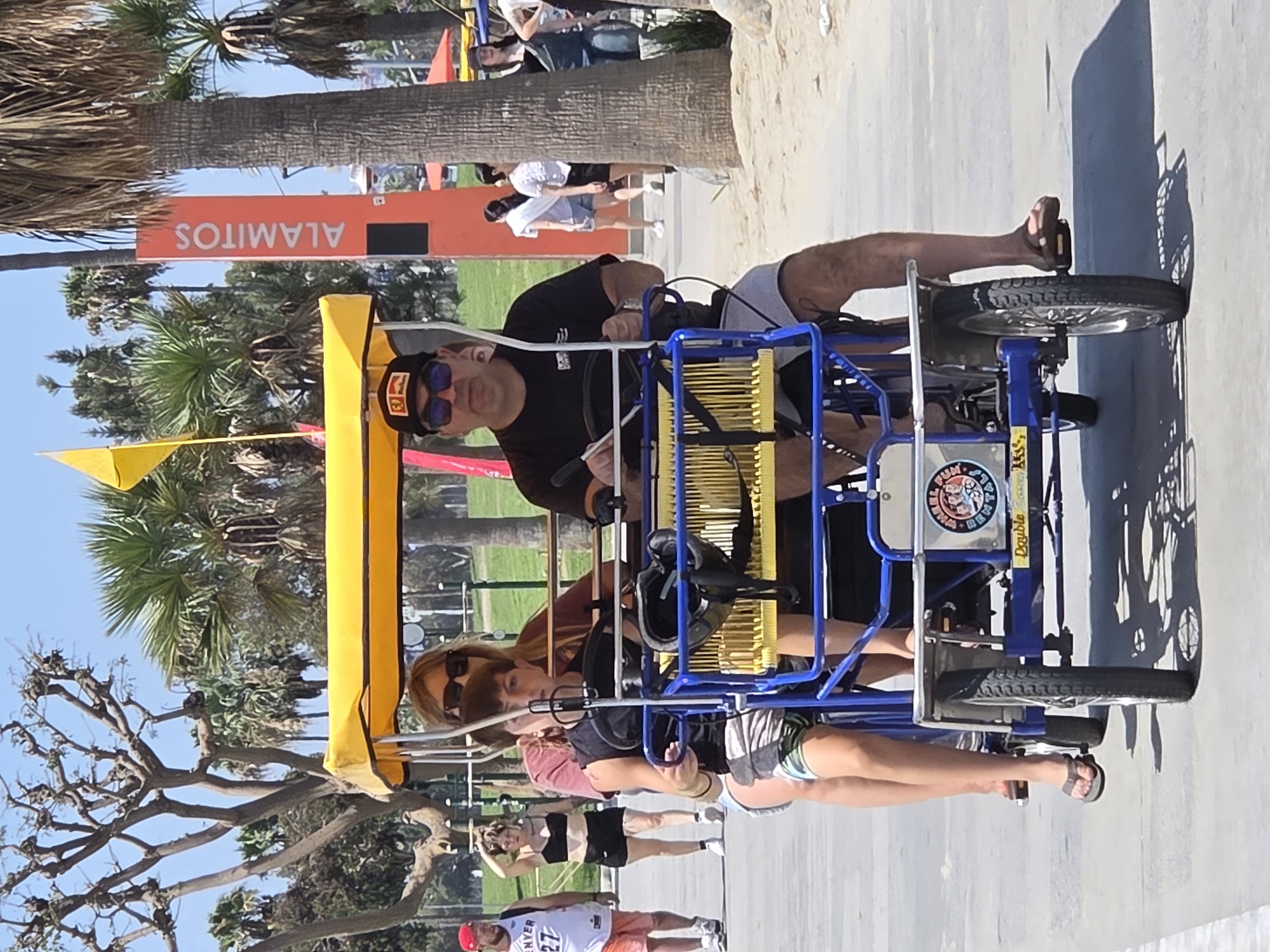 In Long Beach, California, on a sunny day, four individuals are riding a blue and yellow multi-person bicycle on a paved path. The bicycle has a yellow canopy, and the people are seated in two rows. The front rider is a man wearing sunglasses and a black t-shirt, with a black baseball cap. To his right, slightly behind, is a woman in sunglasses and a maroon shirt. In the second row, a young person in a black t-shirt and patterned shorts is seated, and to their right, partially obscured, is another individual. The background features palm trees, a tall orange sign that reads "ALAMITOS," and other people walking and standing. The environment appears to be a beachside boardwalk or park area.