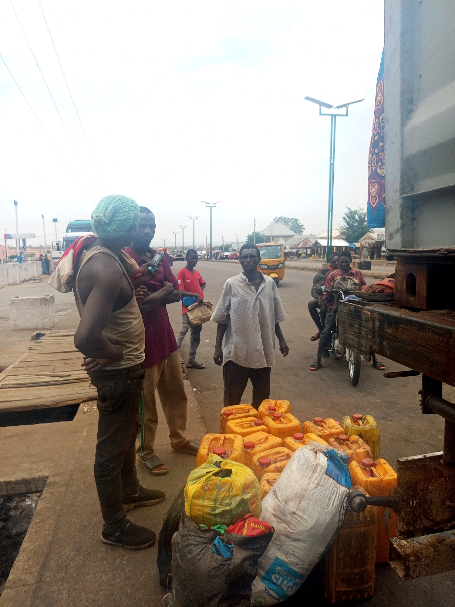 A street scene in Gombe, Nigeria, presents a group of men gathered around a collection of yellow plastic jerrycans, bags, and a portion of a truck. The foreground shows several men standing near the pile of jerrycans and bags, which include a sack labeled "SUGAR". Further back, the image reveals additional people on a motorcycle and in the vicinity, along with a "keke" or auto-rickshaw. The background includes buildings and street lighting. The sky is overcast.