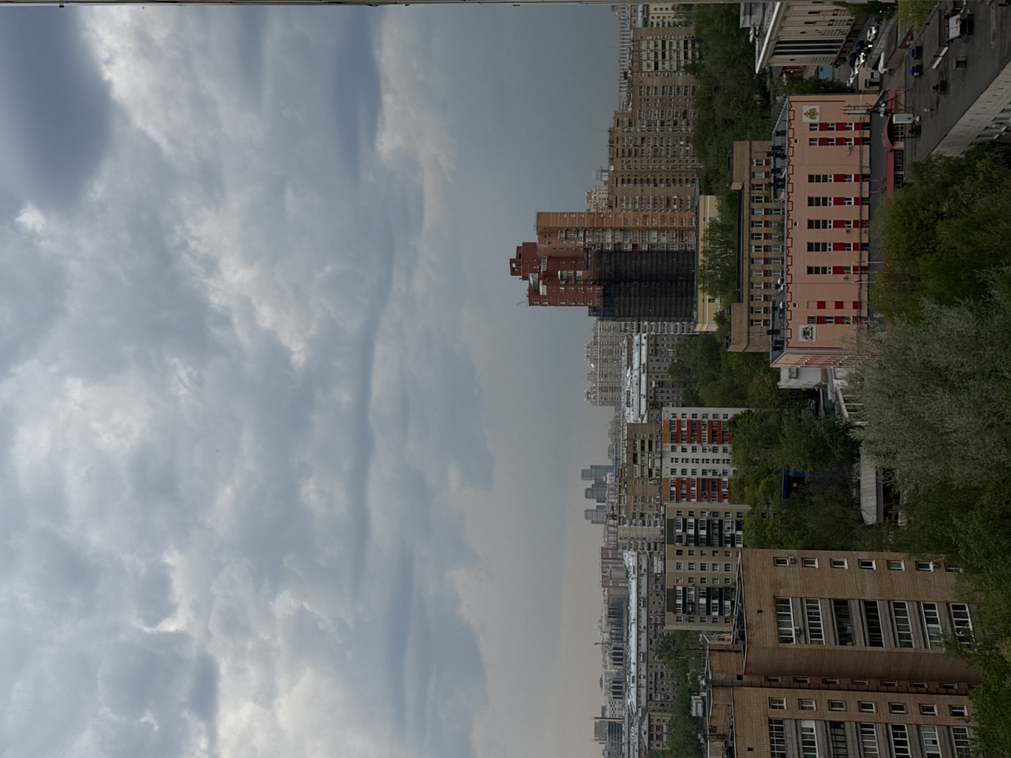 The image is a low-angle, medium shot of a cityscape, with a significant portion of the sky visible. The city appears to be Moscow, Russia, judging from the architectural style of the buildings.
The sky is filled with clouds, varying in color from light gray to dark gray, which suggests overcast conditions. The city comprises numerous buildings of different heights and designs, including residential blocks. Some buildings feature rows of windows and balconies, while others have more ornate facades. Green trees are interspersed throughout the city, possibly indicating parks or residential areas with greenery. A few cars are visible on roads in the distance, adding to the urban landscape.
