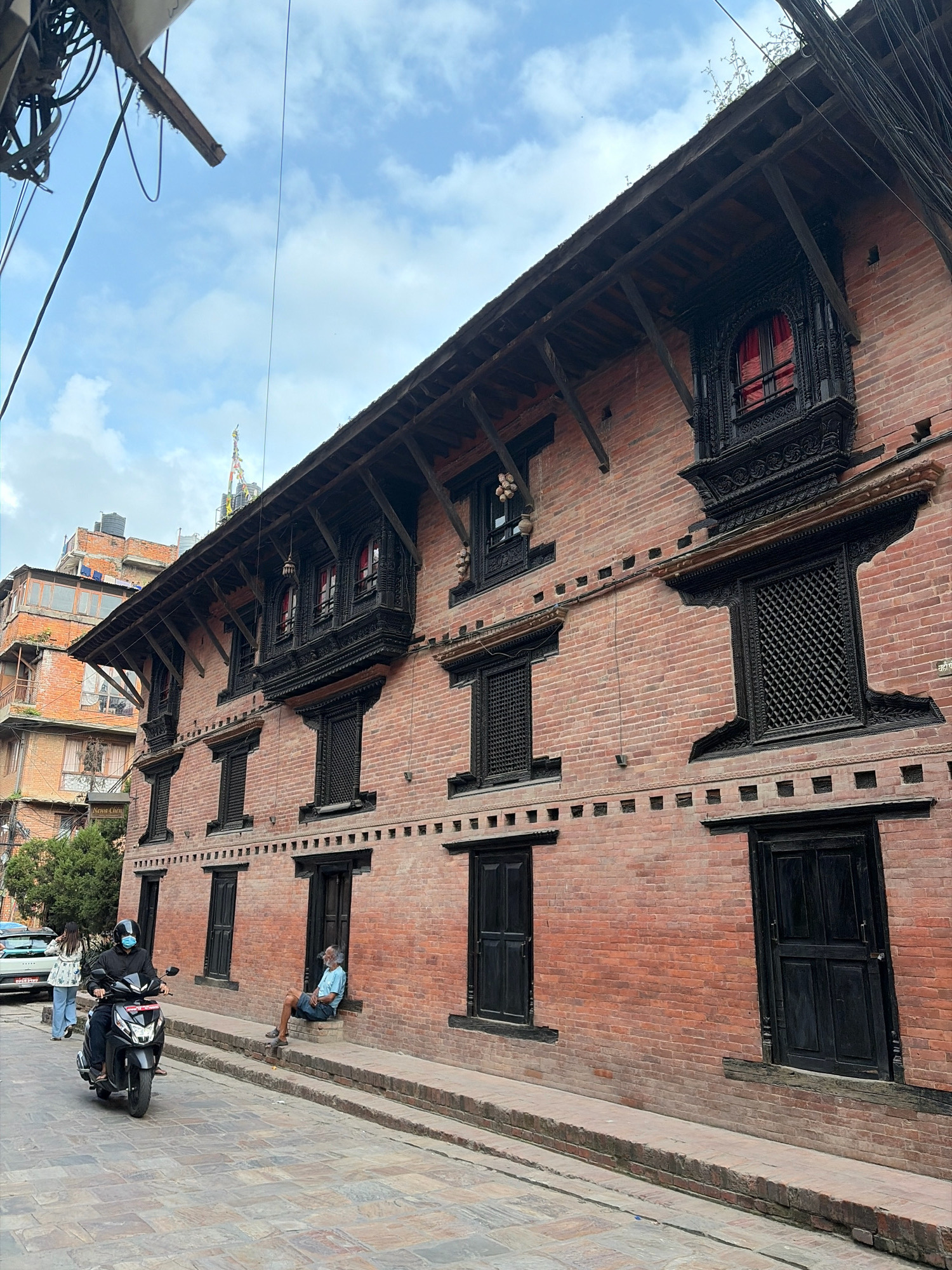 This image depicts a street scene in Lalitpur, Nepal, dominated by a traditional brick building with intricately carved wooden windows. The building's facade features multiple stories with ornate, dark-colored window frames, some of which have red curtains visible inside. The roofline has a prominent wooden overhang supported by diagonal beams. In the foreground, a paved street curves gently. A person on a black scooter, wearing a helmet and face mask, is riding past. To the left of the scooter, a woman in a light-colored jacket and blue jeans walks away from the camera. Further down the street, a man in a light blue shirt and dark shorts sits on a step outside the building, smoking. In the background, another brick building with balconies and water tanks is visible, as well as some greenery. Overhead, electrical wires are strung across the sky, which is partly cloudy. The architecture suggests a historical or culturally significant area within the city.