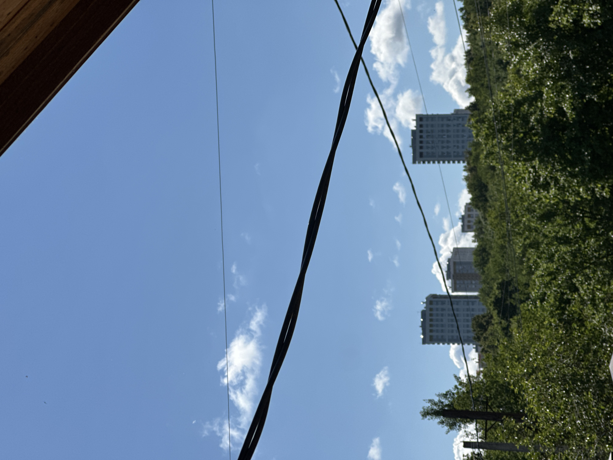 The image presents a bright, sunny daytime scene over what appears to be a residential area in Nizhny Novgorod, Russia. The sky, a vibrant clear blue, is dotted with scattered white cumulus clouds.
In the upper left corner, a dark wooden beam or architectural element suggests the photo was taken from beneath a structure, possibly a roof overhang. The foreground and midground are crisscrossed by multiple dark power lines; a prominent, thick twisted cable runs diagonally from the upper-left toward the lower-right, while a thinner, straight cable is visible further to the left.
Below the expanse of sky, a dense line of lush green trees forms a verdant backdrop, obscuring much of the lower half of the image. Above the tree line, several tall, multi-story buildings, likely apartment blocks, are visible. These structures appear light-colored, with numerous windows, and are primarily concentrated on the right side of the frame. A wooden utility pole with attached wires is partially visible behind some trees in the lower right portion of the image.
The scene captures a typical urban-natural interface, with no specific activity or people visible. The bright lighting and clear sky indicate a pleasant day. There is no visible text within the image.