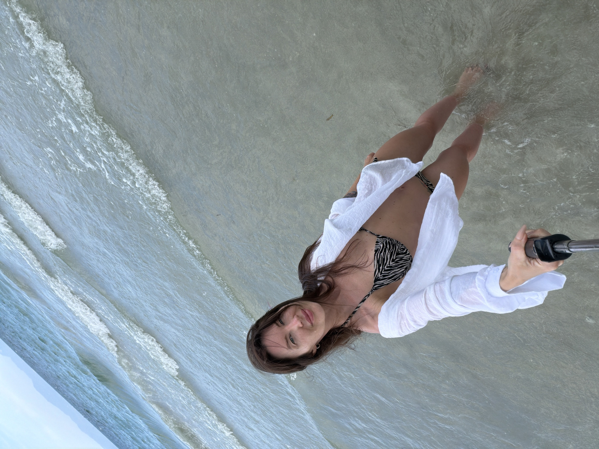 A woman stands in shallow water on a beach, holding a selfie stick. She is wearing a white open shirt over a black and white patterned bikini. The water is clear and calm, with small waves breaking near the shore. The background shows the ocean and a hazy sky.