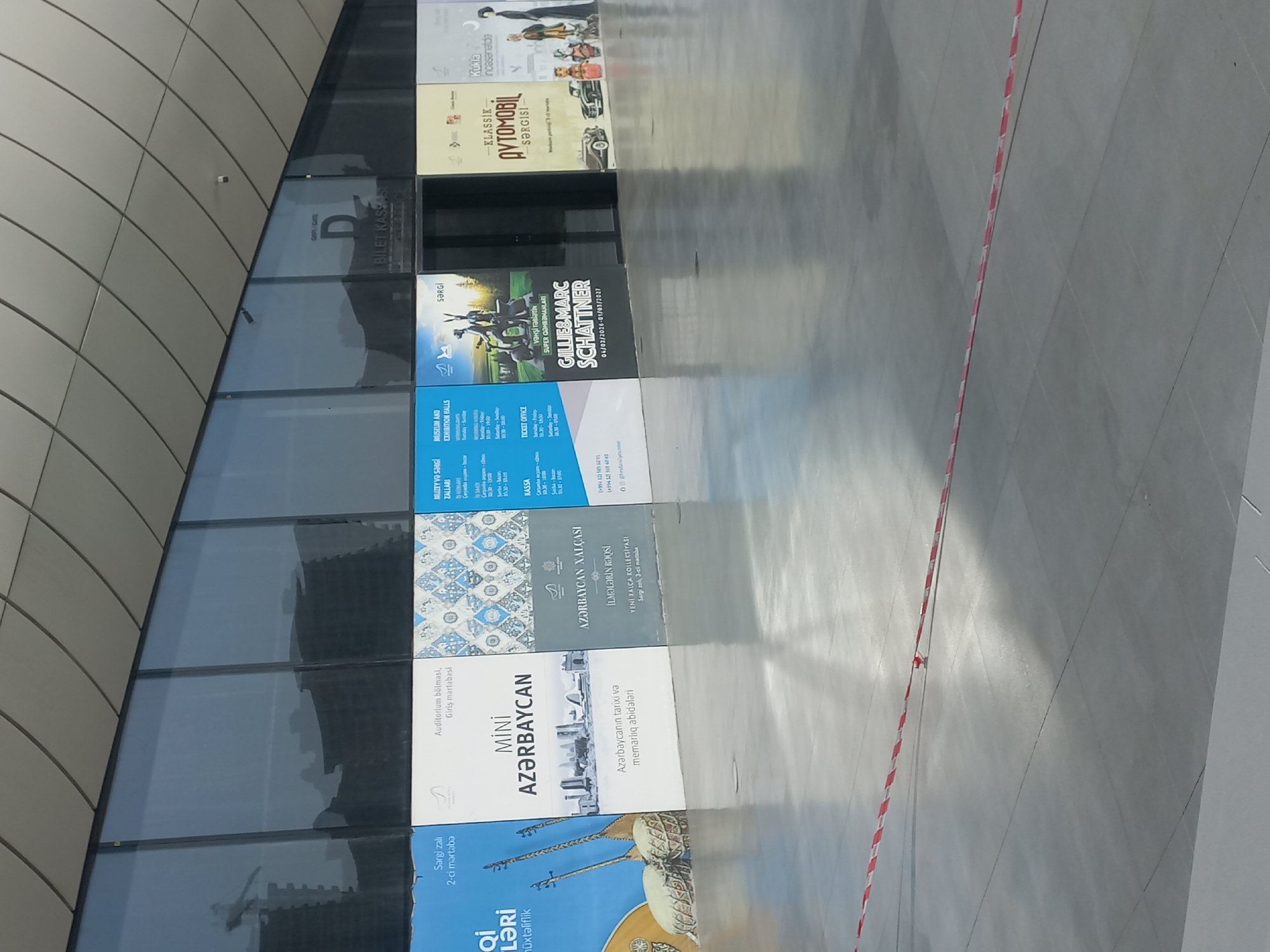 The image shows a row of large posters displayed on the exterior of a modern building with a glass facade. The posters advertise various exhibitions and events, including "MINI AZERBAIJAN," "AZERBAIJAN XALÇASI İLMƏLƏRİN RƏQSİ" (Azerbaijan Carpet Dance of Patterns), "KLASİK AVTOMOBİL SƏRGİSİ" (Classic Car Exhibition), and "Kukla İncəsənətdə" (Puppets in Art). The text on the posters suggests that this location is likely a museum or exhibition center in Baku, Azerbaijan, given the Azerbaijani language used. One poster mentions "AZƏRBAYCAN" and shows images of Azerbaijani cultural elements, further reinforcing the location. The ground in front of the posters is a smooth, polished concrete floor, and a red and white striped caution tape is visible in the foreground, indicating a restricted area. The building's architecture features a curved, modern roof with diagonal supports. Reflections of buildings and cranes are visible on the glass facade.
