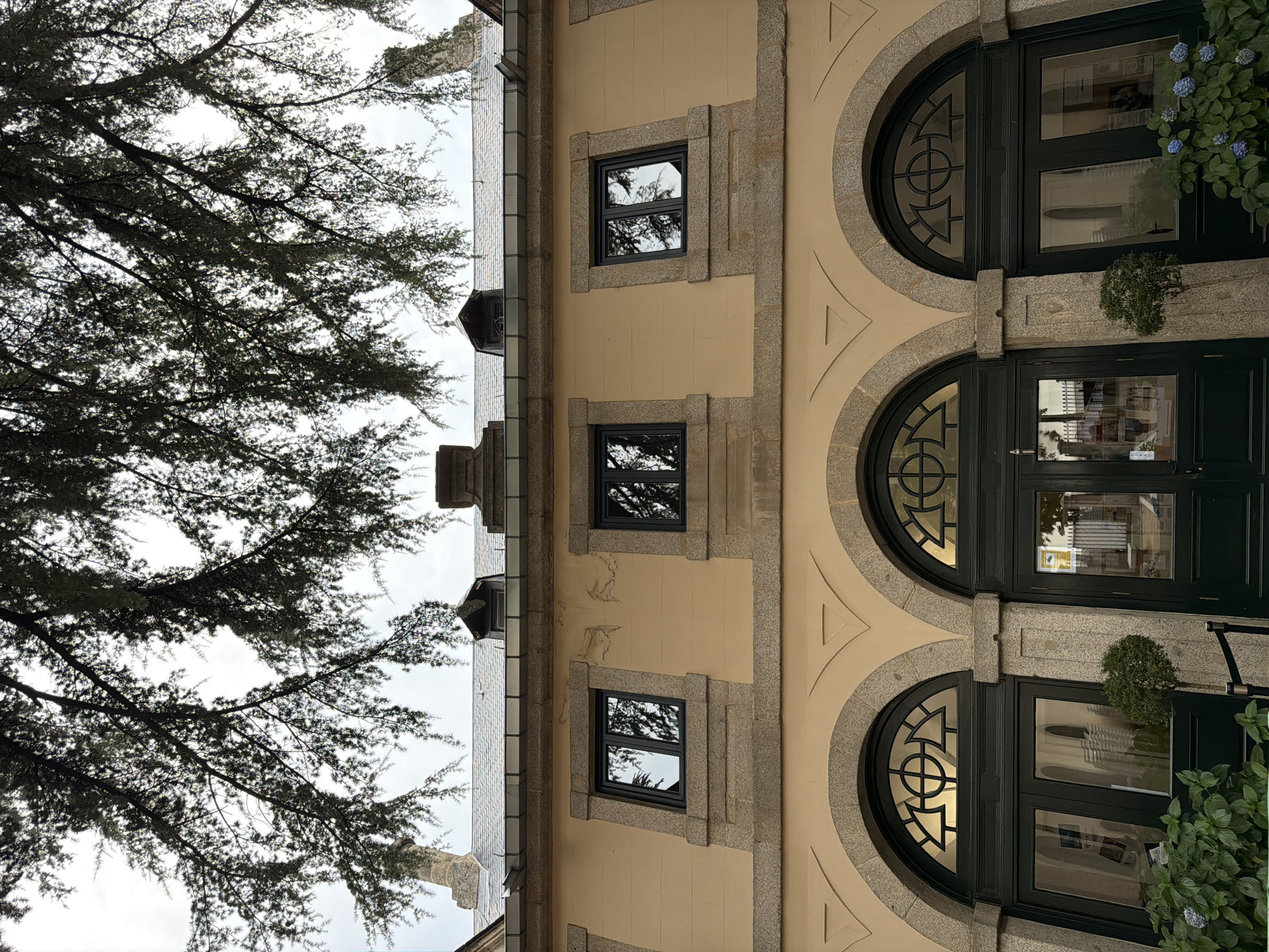 The image shows the facade of a building with an ochre-colored stucco exterior, stone trim, and dark green doors. Three arched entrances are visible at the bottom, each with large glass doors and decorative metalwork above. Above the entrances, there is a band of windows, each with a dark frame and stone sill. The windows reflect the surrounding trees. The roof is tiled and slopes downward, with several dormer windows and chimney structures. The branches of a large tree with dark needles overhang the top of the building, obscuring a portion of the sky. The scene is an exterior view of a building located in San Lorenzo de El Escorial, Spain.