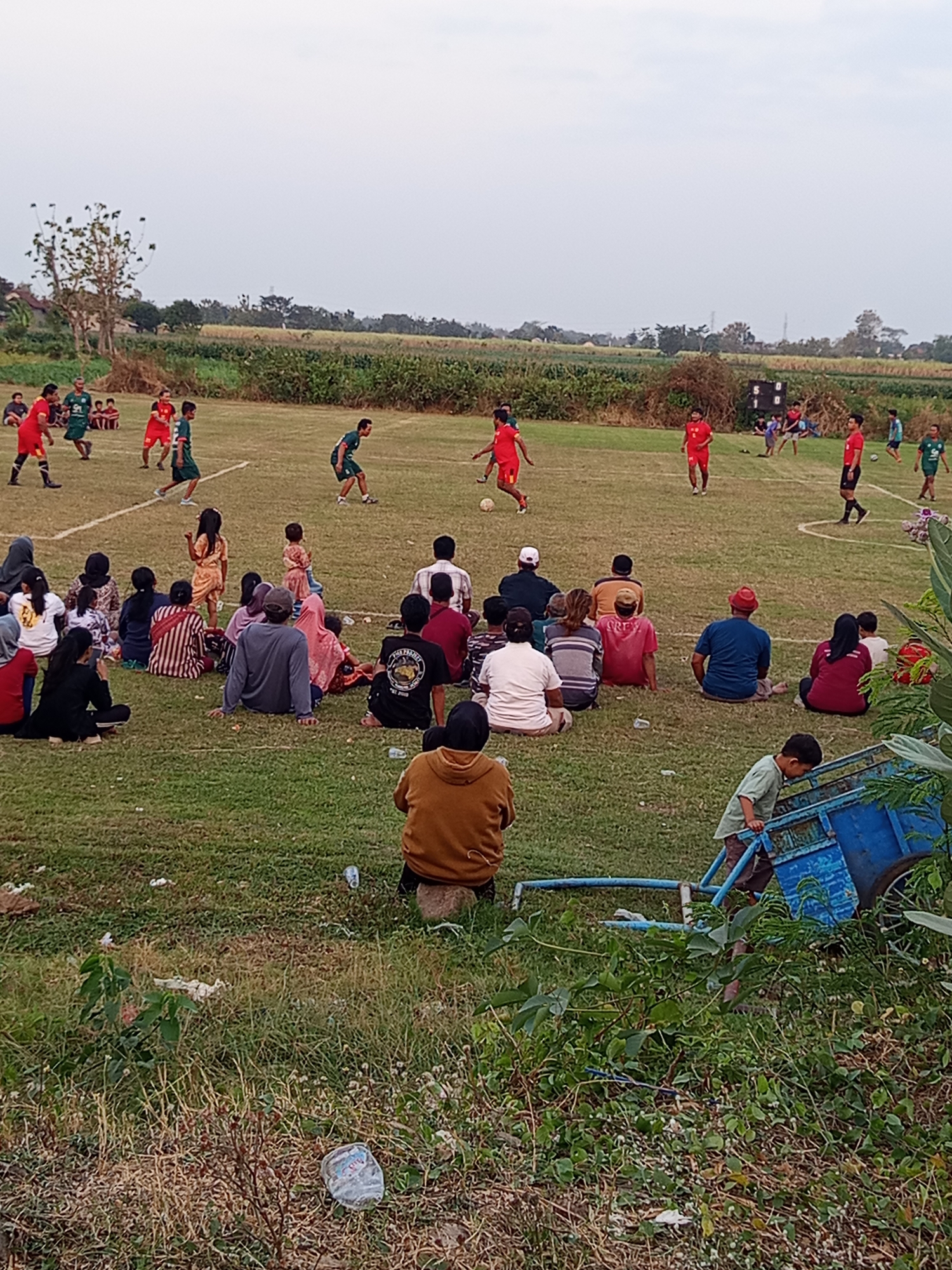 The image depicts a soccer game taking place on a grassy field, likely in Mrican, Indonesia, given the context provided. Several individuals are actively participating in the game, wearing jerseys in red and green. Spectators are seated on the grass around the field, observing the match. Among the onlookers are children and adults, some dressed in traditional attire. The environment appears to be rural, with agricultural fields and trees visible in the background. A scoreboard with visible numbers suggests the game is in progress. A blue cart is positioned on the grass near the spectators. The overall scene conveys a community gathering centered around a sporting event.