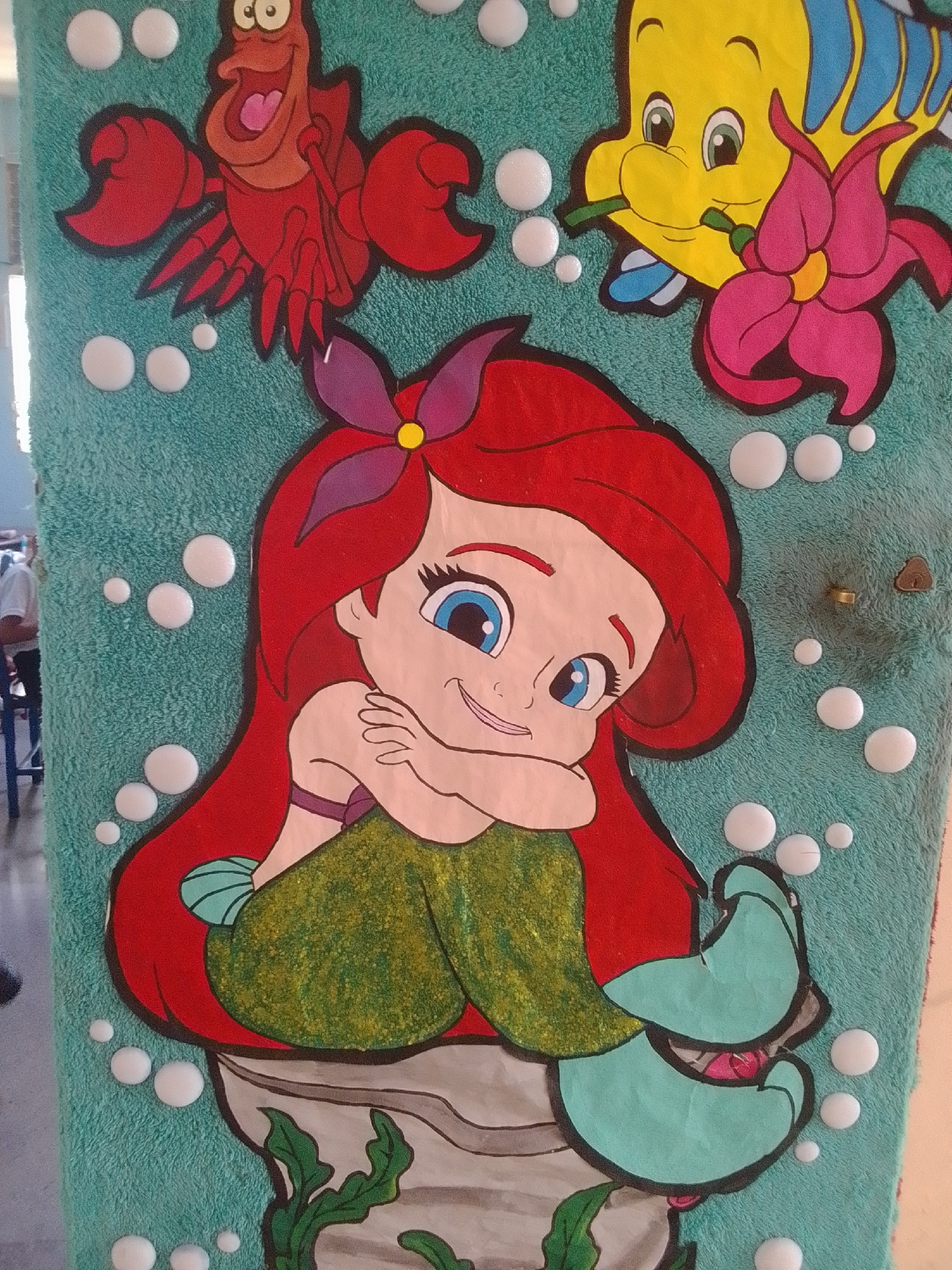 This is a close-up, vertical, full-frame photograph of a wall decoration featuring characters from Disney's "The Little Mermaid." The background is a textured, teal-colored fabric, possibly a towel or carpet, dotted with numerous raised, white, circular embellishments resembling bubbles.
In the center and dominant in the frame is a cartoon depiction of Ariel, the mermaid. She has vibrant red hair, large blue eyes, and is smiling. She is wearing a green sequined tail and a purple bow in her hair. Ariel is posed sitting on a rock-like structure with green seaweed.
To the upper left of Ariel is Sebastian, the crab, depicted in bright red with black outlines and an open mouth, appearing to be shouting. To the upper right of Ariel is Flounder, the yellow fish, with blue fins and a pink flower held in his mouth.
The overall scene is playful and colorful, characteristic of children's decor. The image does not provide any specific environmental context within La Tagua, Venezuela, beyond the fact that it is a decorative element likely found in a public or private space. There are no identifiable people in the immediate foreground or interacting with the artwork.