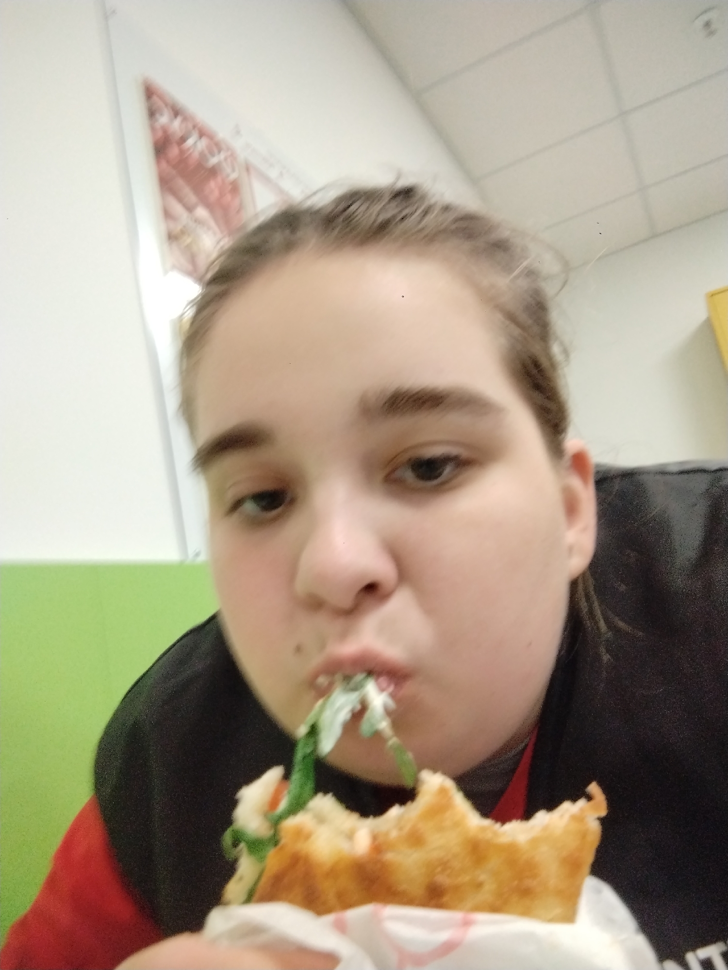 A young individual, possibly a teenager, with light skin and brown hair pulled back, is captured in a candid moment eating a large sandwich indoors in Plovdiv, Bulgaria. They are seen mid-bite, with green leafy vegetables, likely arugula, protruding from their mouth and the sandwich. The food item appears to be a thick, possibly focaccia-style bread filled with white sauce or cheese and the green leaves, wrapped in white paper. The person is wearing a black top over a red undergarment.
The setting is an indoor space, possibly a cafeteria, school, or casual eatery. The background features a white drop ceiling with a grid pattern and white walls. A prominent bright green horizontal panel is visible on the lower left wall, and a blurry, light-colored poster with a red/pink triangular motif is on the upper left wall. A small yellow object or cabinet is partially visible on the right. The lighting is bright and even, consistent with an indoor environment. The individual's expression is one of concentration on eating. There is no discernible text visible in the image.