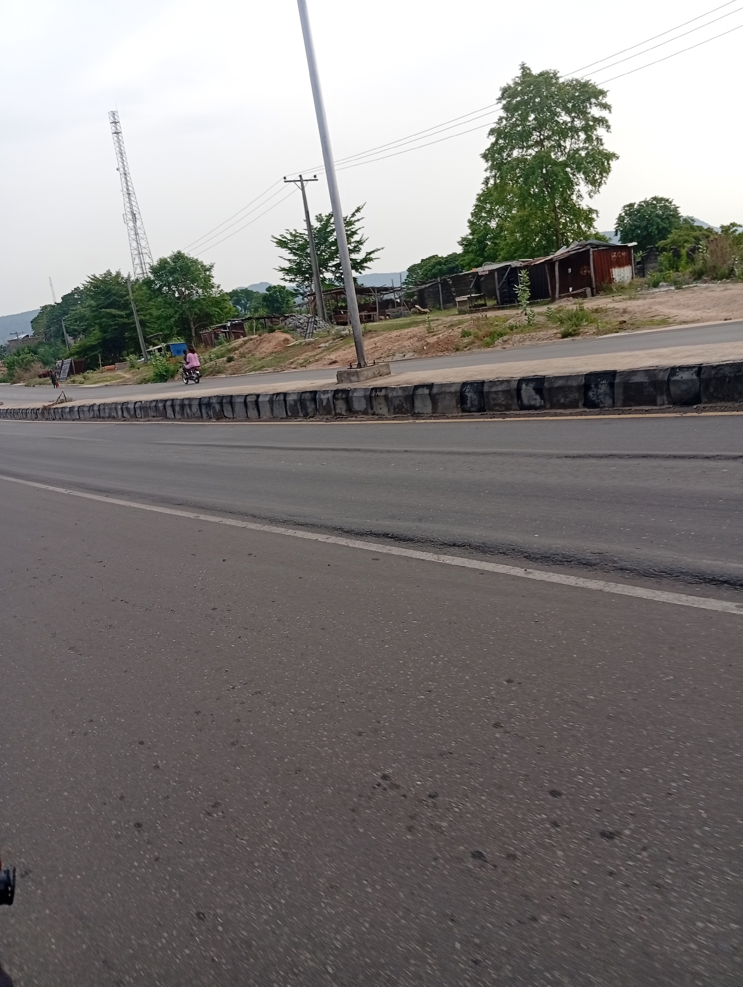 The image shows a section of a road in Lokoja, Nigeria. The road is paved and has a white line marking the lane. A concrete barrier separates the road from the shoulder. There is a person riding a motorcycle on the shoulder of the road. In the background, there is a communications tower, trees, and some buildings that appear to be simple structures. Power lines run along the roadside. The sky is overcast.