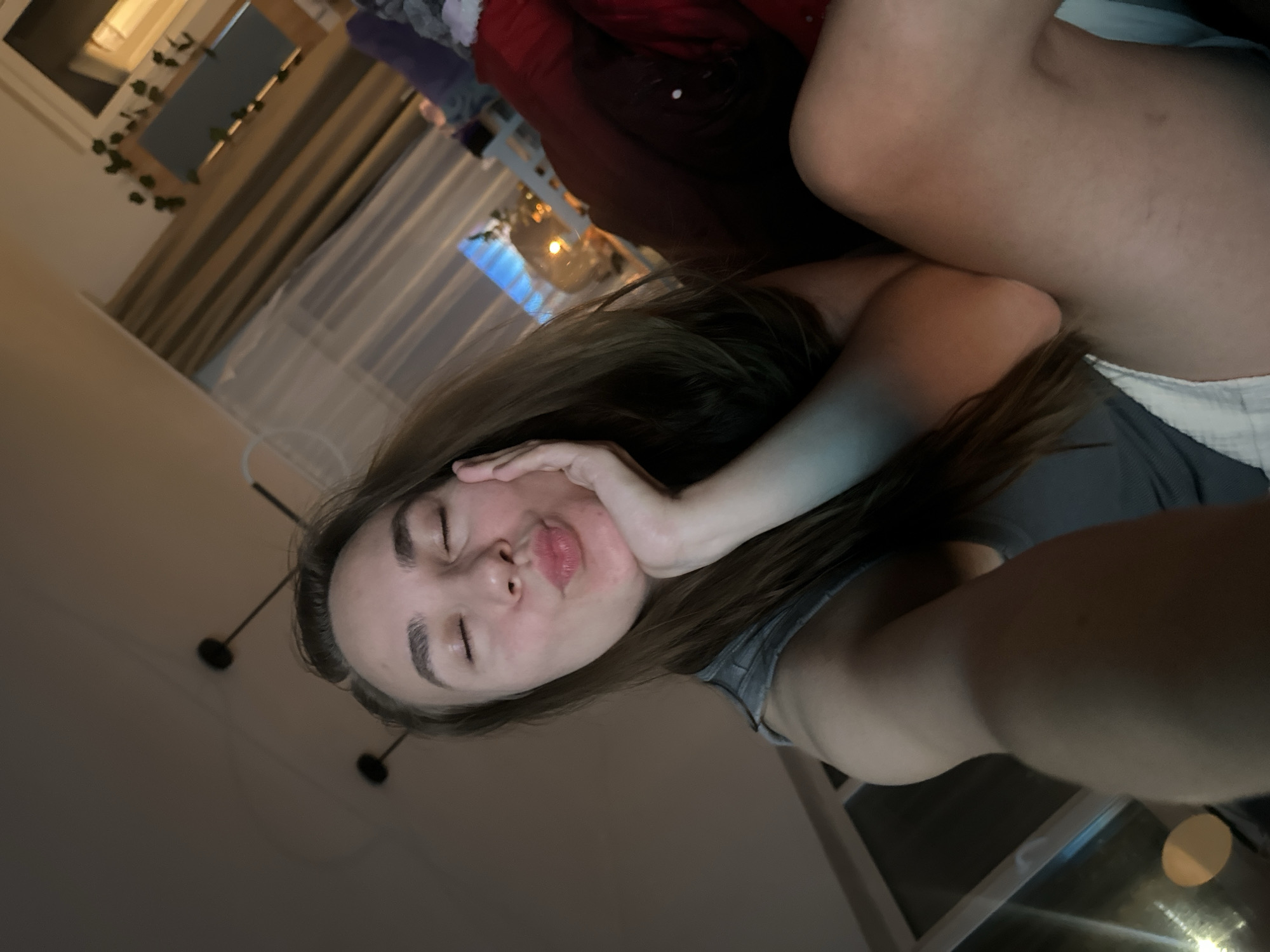 A woman is lying down, face directed towards the camera, with her eyes closed and lips pursed. Her right hand is against her cheek. She has long brown hair and is wearing a grey tank top. She appears to be on a bed with a red blanket. In the background, there is a window with curtains and string lights, and a modern hanging light fixture. The location in the city of Kharnavy, Russia is not possible to determine from the image.