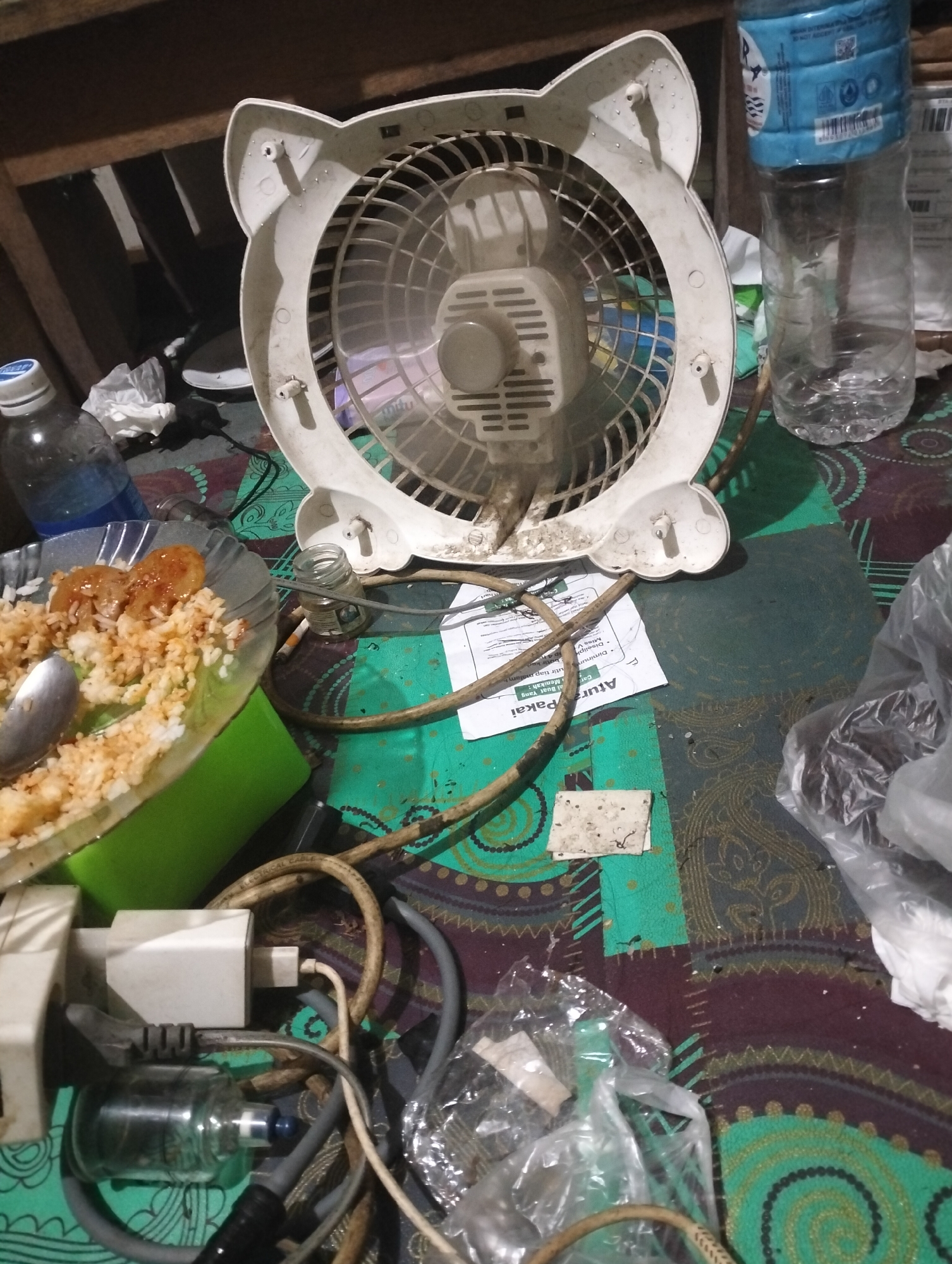The image depicts a cluttered tabletop scene, likely in a residential setting in Mandala Jaya, Indonesia. In the foreground, an electric fan with a white, cat-ear-shaped casing sits prominently, showing signs of dust accumulation. To the left of the fan, a plate of rice with what appears to be meat or sauce is partially visible, accompanied by a spoon. Several power cords and adapters are strewn across the surface, connected to a white electrical component in the lower left corner. A clear glass bottle, possibly containing a liquid, is also present. Behind the fan, a partially full plastic bottle of water is standing upright. A crumpled piece of paper with Indonesian text, possibly instructions or a label, is lying near the fan. Various other objects, including a plastic bag and a lit cigarette butt, are scattered around the scene, contributing to a general sense of disarray. The overall lighting suggests an indoor environment, possibly at night, with artificial light sources.