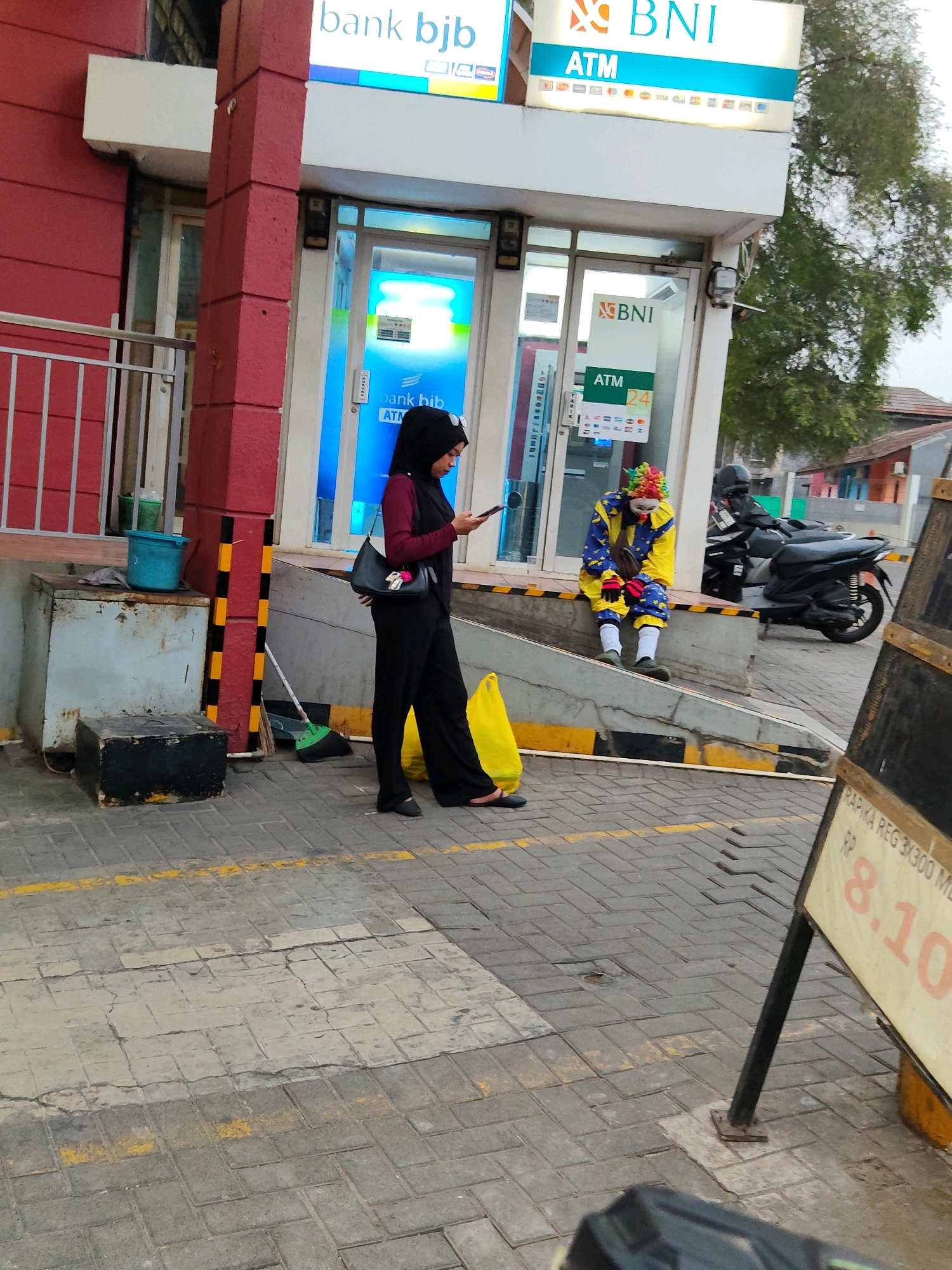 The image captures an outdoor scene in front of an ATM kiosk, likely located in Purwakarta, Indonesia. A woman wearing a black hijab and dark clothing is standing and looking at her phone. To her right, a person in a clown costume, with a colorful wig, painted face, and yellow and blue outfit, is sitting on a low wall. Behind them, a motorcycle is parked. The ATM kiosk has branding for "bank bjb" and "BNI," indicating these are the financial institutions. The ground is paved with interlocking bricks, and yellow lines are painted on the pavement. In the foreground, a sign with text including "RP 8.10" is visible. The overall environment suggests a public space, possibly near a street or commercial area.