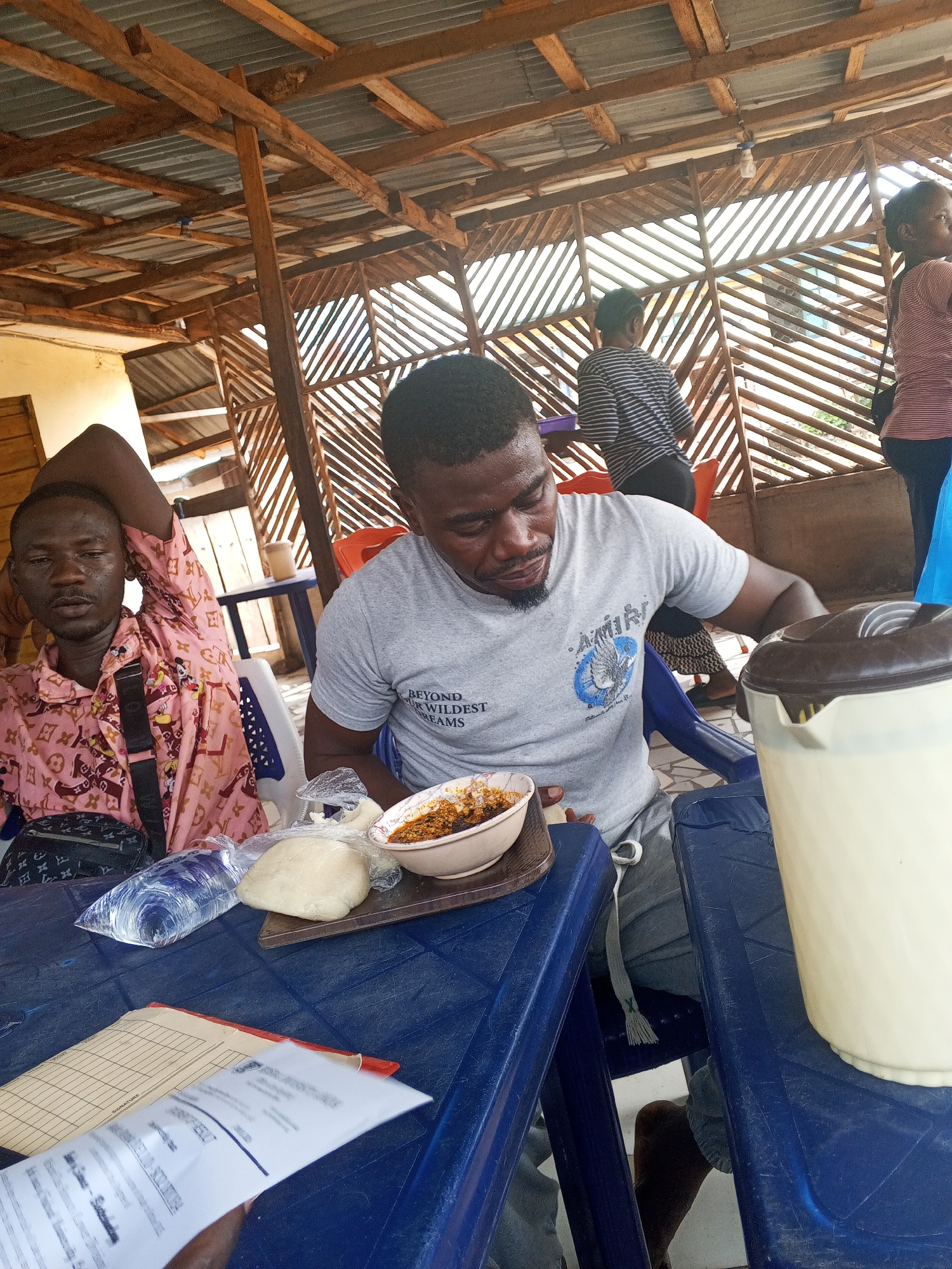 In Lokoja, Nigeria, two men are seated at a blue plastic table under a rustic, open-air shelter with a corrugated metal roof supported by wooden beams. The man on the left wears a pink shirt with a repeating pattern of the Louis Vuitton logo and a black crossbody bag with a similar pattern. He is resting his head on his raised left arm. The man on the right wears a grey t-shirt with a graphic and text reading "BEYOND OUR WILDEST DREAMS." In front of them on the table are two bowls of food, one containing a stew-like dish, and a large serving of fufu, a staple food in West Africa, wrapped in plastic. A large, beige plastic water dispenser is also visible on the table. In the background, other individuals, including two women, are present, and the shelter's walls are constructed with diagonal wooden slats. A document, possibly a form or receipt, is on the table in the foreground.