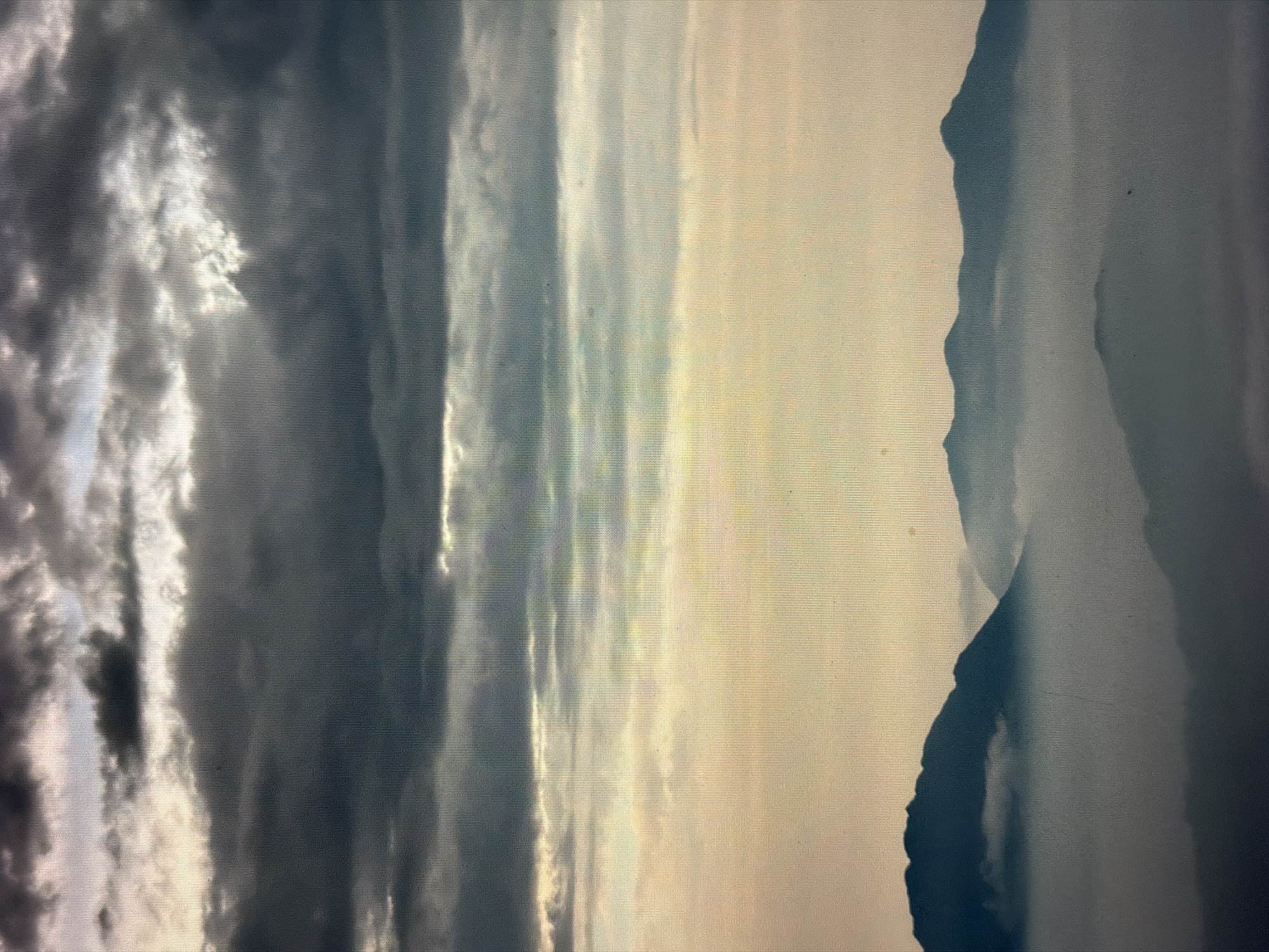 The image presents a landscape consisting of clouds, sky, and mountainous terrain. Dark, turbulent clouds dominate the left side of the frame, contrasting with the brighter, ethereal sky in the center. The right side of the image features a mountain range, silhouetted against the sky. The exact location in Đà Nẵng is indeterminable from the image alone. The image contains no people.