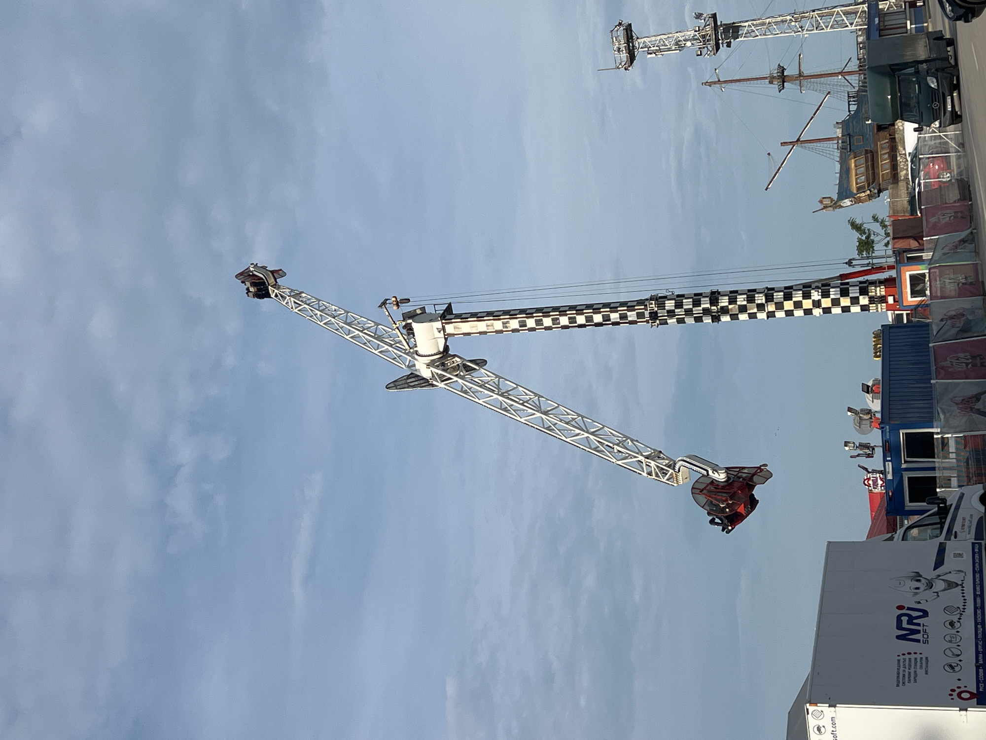 This image depicts an active outdoor amusement park or carnival scene during daylight hours under a clear, light blue sky with minimal cloud cover. The central subject is a large pendulum amusement ride, characterized by its white truss structure and a prominent black and white checkered support pillar. A red gondola, containing people, is visible at the end of the pendulum arm, positioned high against the sky, indicating the ride is either in motion or at its peak.
In the background, another tall, white tower ride stands, and a large, ornate pirate ship facade adds to the themed entertainment setting. Various other structures, possibly stalls or tents, are partially visible on the right side of the image. A white commercial truck is parked in the foreground, displaying branding and information.
Visible text on the side of the truck includes "NRI SOFT" and "soft.com". Below this, there is Bulgarian text: "ВИДЕОНАБЛЮДЕНИЕ" (Video Surveillance), "СИСТЕМИ ЗА ДОСТЪП" (Access Systems), "ПАРИЧНИ РЕШЕНИЯ" (Payment Solutions), "ЗАРЕДНИ СТАНЦИИ" (Charging Stations), and "МОНТАЖ ИНСТАЛАЦИЯ" (Installation). Additionally, a list of Bulgarian cities is present: "РУСЕ - СОФИЯ - ВАРНА - БУРГАС - ПЛОВДИВ - ХАСКОВО - ПЛЕВЕН - ВЕЛИКО ТЪРНОВО - СТАРА ЗАГОРА", confirming the location context of Bulgaria, specifically Varna. The scene conveys a sense of excitement and entertainment typically associated with amusement parks.