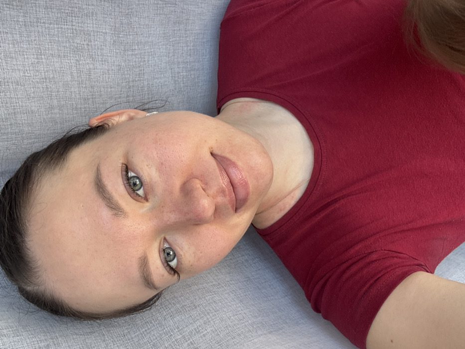 An adult female is depicted in a close-up, selfie-style indoor portrait, positioned diagonally, looking directly at the viewer with a slight, closed-lip smile. She has light skin with subtle freckles across her nose and cheeks, green-blue eyes enhanced with what appear to be eyelash extensions, and light brown eyebrows. Her dark hair is pulled back from her face. A small, silver-colored hoop earring is visible in her right ear. She wears a plain, short-sleeved, maroon crew-neck t-shirt. A faint gold-colored chain is visible beneath her collar. Minor red skin irritations are present on her neck and upper chest. In the lower right corner, a partial view of light-colored hair, consistent with that of a child, is discernible. The background is a uniform, light gray textured surface, possibly fabric. The scene is an unidentified indoor location; no visual information indicates the city of Shul'gino, Russia.