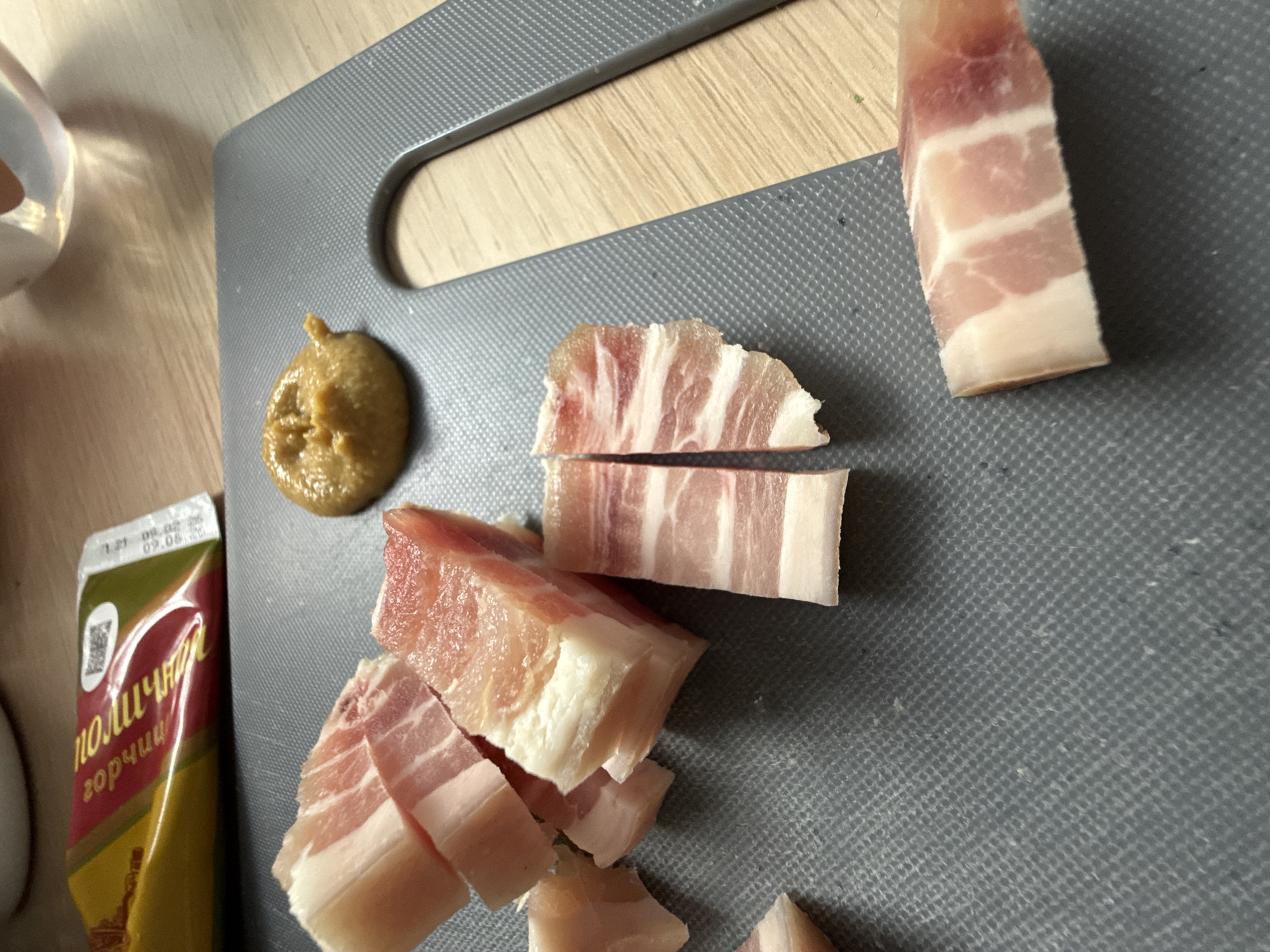 This close-up image captures a culinary scene on a grey cutting board, situated on a light wooden surface, likely a kitchen counter in Moscow, Russia. The primary subjects are slices of raw or cured pork belly (salo) and mustard.
To the right, a long, rectangular piece of pork belly exhibits distinct horizontal layers of pink meat and white fat. More irregularly cut pieces of pork belly, varying in size from chunks to smaller slices, are scattered towards the center and left of the cutting board, all displaying similar lean-fat marbling.
A dollop of coarse-grain, brownish-yellow mustard is centrally placed on the cutting board, slightly to the upper left of the pork. A soft, yellow and red/maroon mustard pouch is partially visible on the wooden surface to the far left. The Cyrillic text "горчица" (gorchitsa), meaning mustard, is clearly legible on the pouch, along with other Cyrillic characters such as "поличн" which likely indicate a brand or type. A date or code "1.21 08.02.26" and "09.08." is visible on the packaging, along with a QR code.
The scene suggests the preparation of a meal or snack. A clear glass or cup is partially visible on the far left edge of the frame. The lighting indicates an indoor setting. No people are present, and no specific time of day or weather can be determined from the image. The Cyrillic text on the mustard packaging strongly cues the geographic location as a Slavic-speaking region, consistent with Moscow, Russia.