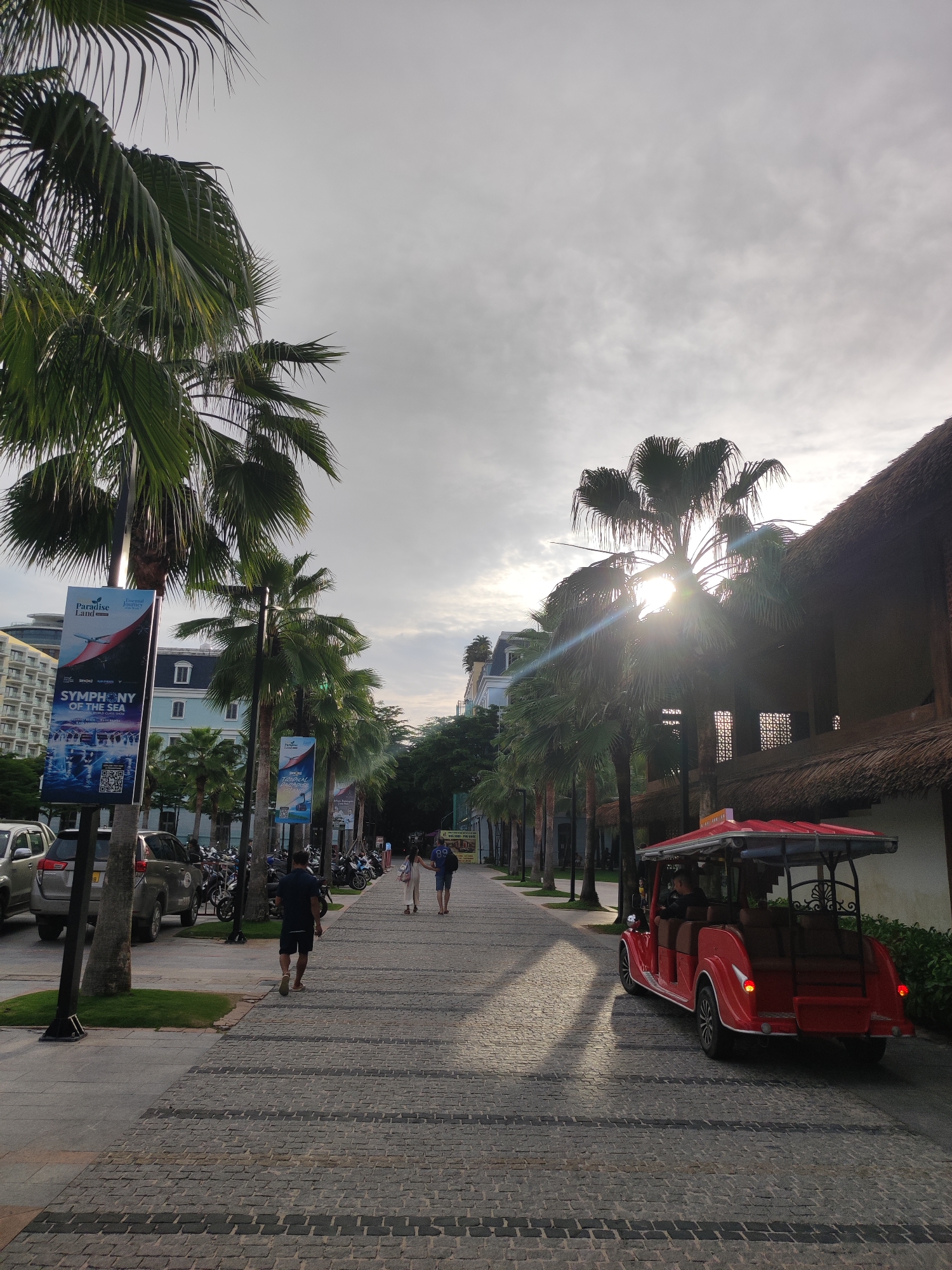 The image depicts a paved walkway lined with palm trees, leading towards buildings with thatched roofs and modern architecture. The sky is overcast with the sun partially visible through the palm fronds, casting dappled shadows on the ground. A red, open-air tourist vehicle is driving away from the viewer on the right side of the walkway. Several people are walking on the walkway in different directions. In the background, there are multi-story buildings and parked motorcycles. A large banner advertising "Paradise Land" and "Symphony of the Sea" is visible on the left side of the walkway. The overall scene suggests a resort or entertainment complex in Phú Quốc, Vietnam, likely during the late afternoon or early evening.