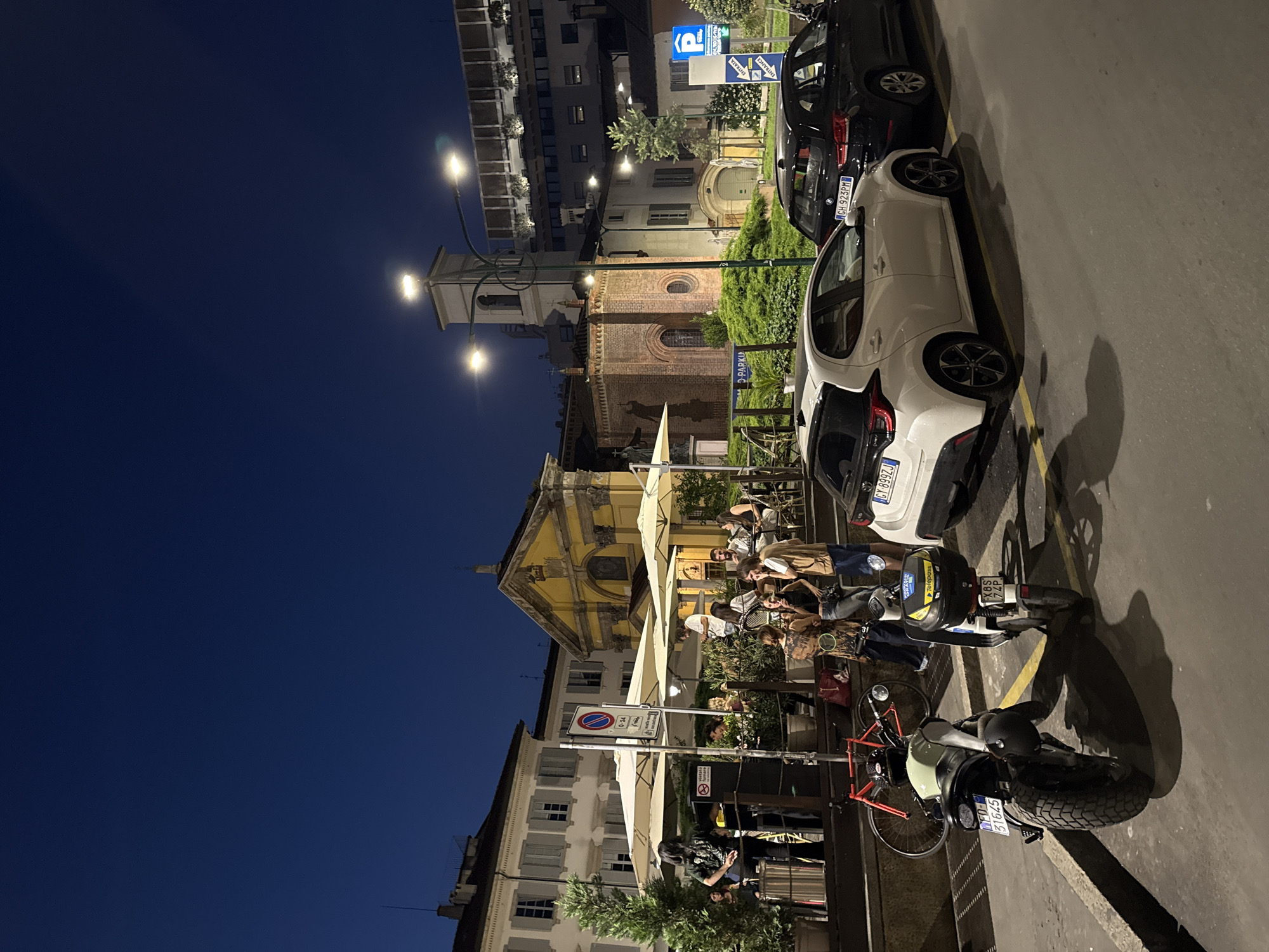 At night in Milan, Italy, a street scene shows outdoor cafe seating with patrons under white umbrellas. Parked cars line the street, including a white Toyota Yaris with license plate "GX 899ZJ" and a black BMW with license plate "GH 975PM." A motorcycle with license plate "FD 51645" and a bicycle are also parked near the cafe. In the background are buildings, including a church and a bell tower illuminated by street lights. The sky is dark blue.