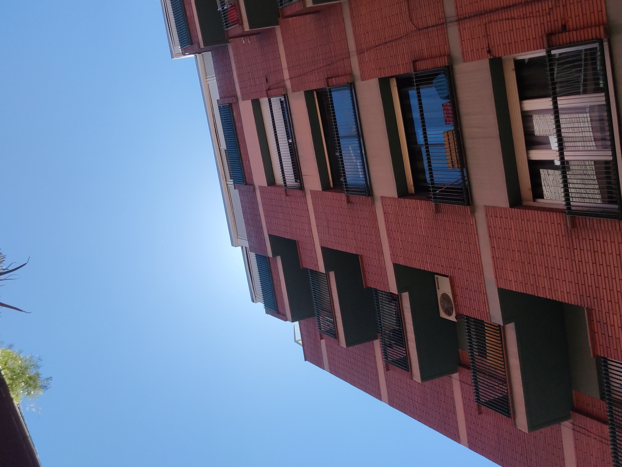 The image shows a low-angle view of a multi-story brick building against a bright blue sky. The building's facade features a repeating pattern of rectangular windows with dark metal grilles. Some windows reflect the sky, while others are open. The building's design includes projecting sections, creating shadowed areas. An air conditioning unit is visible on one of the projecting sections.