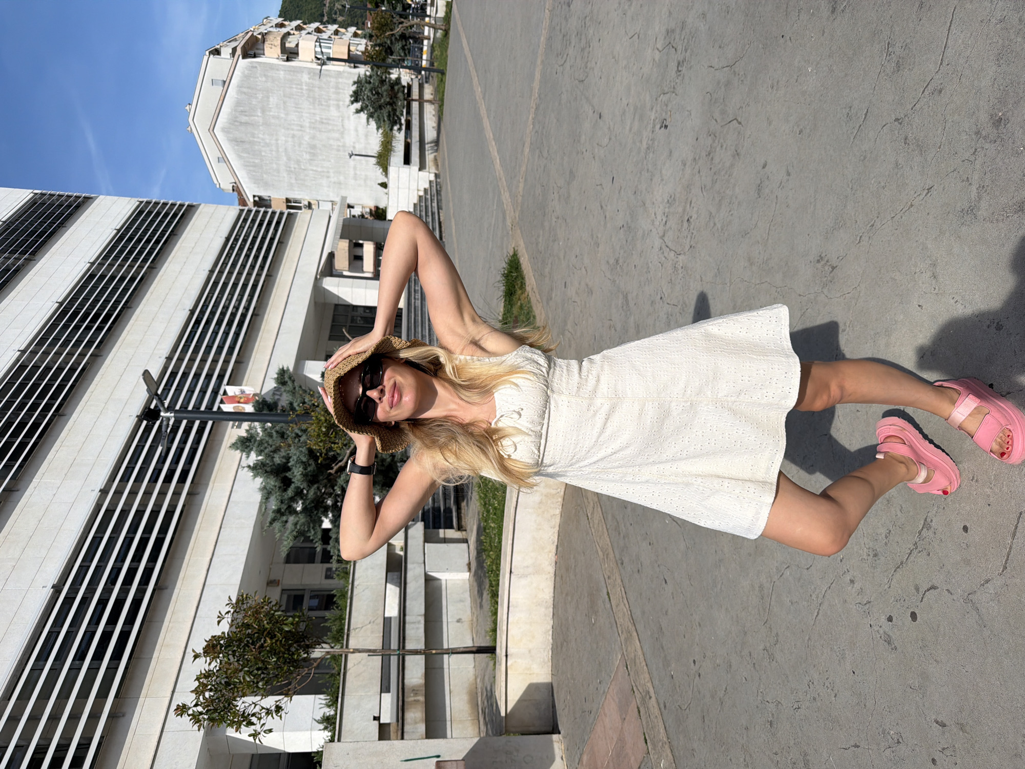 In the city of Budva, Montenegro, a blonde woman in her late twenties stands on a paved public square. She is wearing a short, white eyelet dress, a straw hat, sunglasses, and bright pink platform sandals. Her hands are clasped behind her head, and she is looking towards the camera with a slight smile. Behind her, a modern, multi-story building with horizontal white and dark striped windows dominates the background. Adjacent to it, a lighter-colored building with a peaked roof and balconies is visible. The ground is a textured grey pavement with visible cracks and darker circular stains. Green trees and shrubs are present in the middle ground, and in the distance, a verdant mountainside can be seen. The scene is illuminated by bright sunlight, casting a shadow of the woman on the pavement.