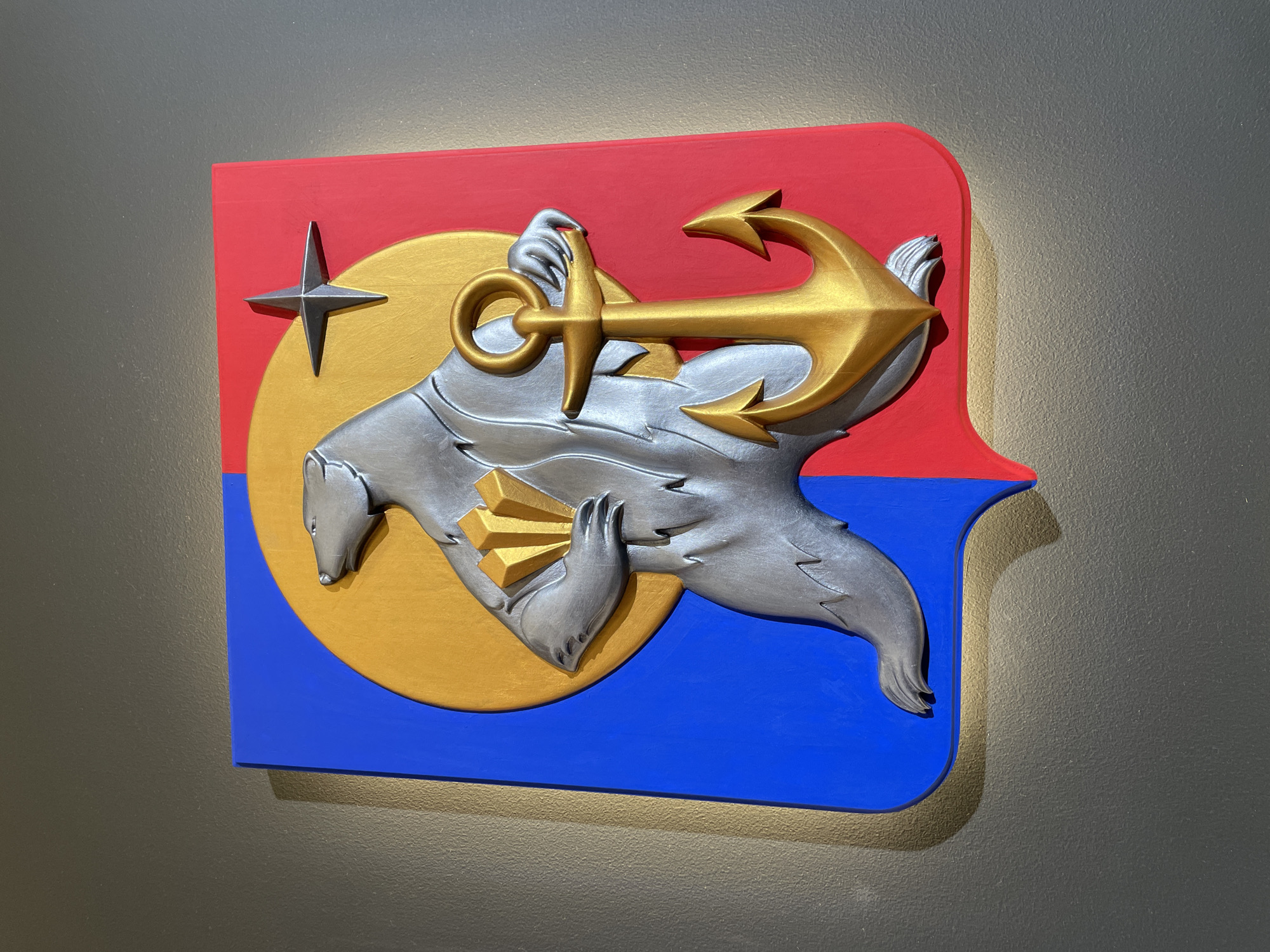 A three-dimensional object or sign hangs on a gray wall. The sign is a red, white, and blue nautical-themed coat of arms. A gold sun is in the middle of the coat of arms. A polar bear sits in front of the sun and holds a gold anchor in its paws. A four-point star is on the upper left. The location is not specified.