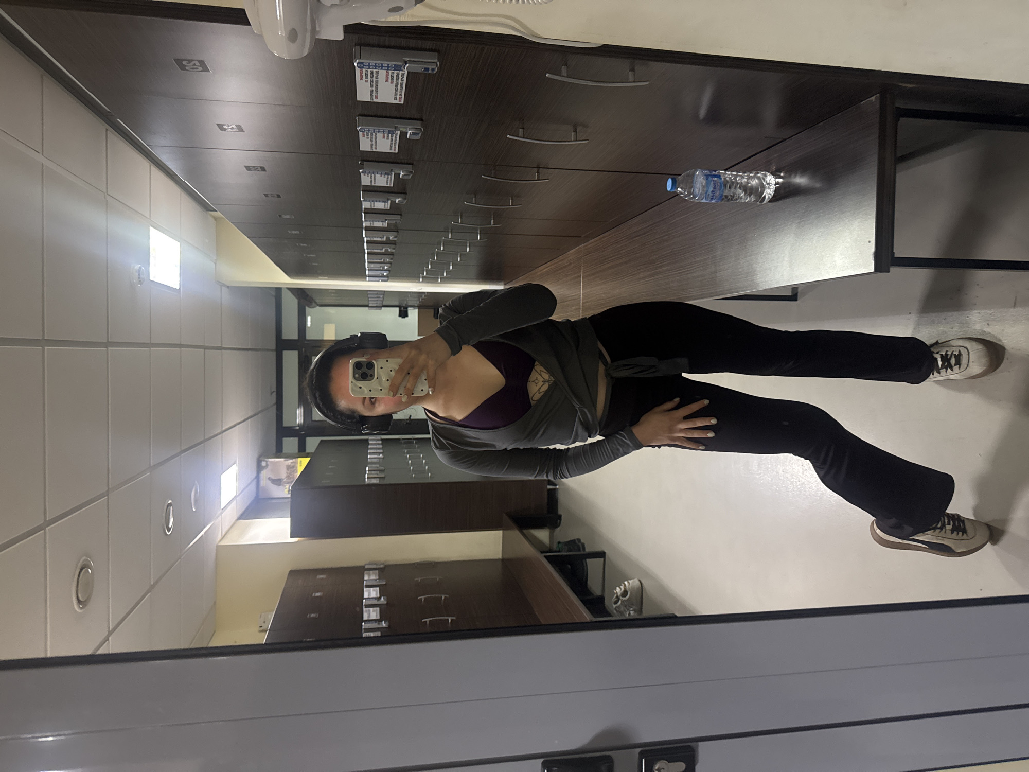 The image is a mirror selfie taken indoors, likely in a locker room or changing area of a gym or sports facility in Burgas, Bulgaria. The original image is rotated 90 degrees counter-clockwise.
The primary subject is a young woman, partially obscured by her phone, taking the selfie. She is wearing dark headphones, a dark purple sports bra or top, a grey long-sleeved wrap-around top, black leggings or flared pants, and light-colored sneakers with dark soles (Puma brand is visible). A tattoo is visible on her left shoulder/chest area. She holds a white phone with a speckled case, which covers most of her face. Her left hand is placed on her hip or waist.
The setting features dark brown wooden or laminate drawer units or lockers with silver handles. Some of these units have small white card holders attached, containing unreadable text. Numbers like "50," "51," "52," and "53" are faintly visible on the upper row of drawer fronts. A plastic water bottle with a blue cap and a label with Cyrillic text (possibly "ДЕВИН," a Bulgarian brand) rests on one of these dark brown surfaces. The walls are light-colored and appear tiled, with square and circular artificial light fixtures. The floor is also light-colored. A white hand dryer is partially visible in the top left corner of the original image.
The activity depicted is the woman taking a mirror selfie, presumably before or after an exercise session. The artificial lighting suggests it could be any time of day.