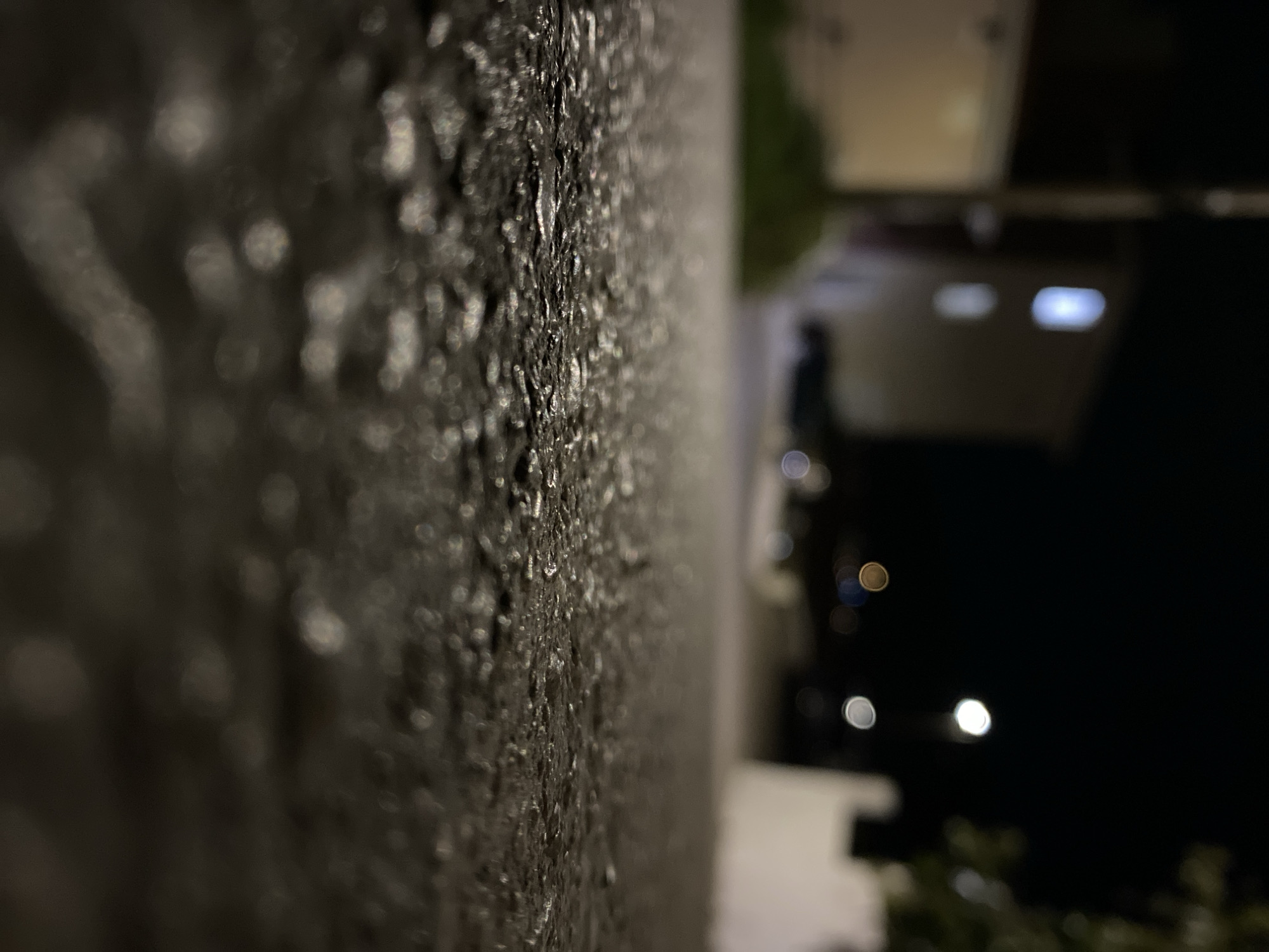 The image presents a close-up view of a textured, possibly wet, grey or dark-colored wall in Budva, Montenegro. The surface is irregular, with what appears to be water droplets or reflective elements scattered across it. In the background, slightly out of focus, is a building or structure with illuminated windows and other light sources. The overall scene is at night, indicated by the darkness surrounding the building in the background. The vantage point suggests an eye-level perspective looking up at the wall. The lack of discernible people or specific actions focuses attention on the texture of the wall and the ambient lighting of the surrounding buildings.