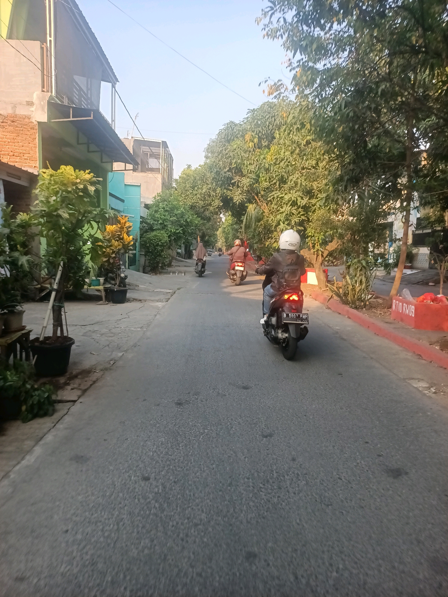 The image depicts a street scene in Telagaasih, Indonesia, characterized by residential buildings and lush vegetation. The road is paved and appears to be a local street, with a slight incline. On the left side of the road, there are several houses with varying architectural styles, some with brick walls and others with more modern facades. Potted plants and small trees are visible along the sidewalk in front of the houses. On the right side of the road, there are mature trees providing shade, and a red-colored curb or planter box is visible in the foreground.
Three individuals on motorcycles are traveling down the street. The closest motorcycle has a rider wearing a white helmet, a dark jacket, and blue jeans. The motorcycle's license plate is visible, reading "B 5687 FHP". Further down the road, two other motorcycles are also in motion. One motorcycle has a rider wearing a red jacket, and another is further ahead with a rider wearing a beige head covering. The overall atmosphere suggests a calm, everyday moment in a residential area of Telagaasih, likely during the late afternoon given the lighting and shadows. The sky is clear and blue.