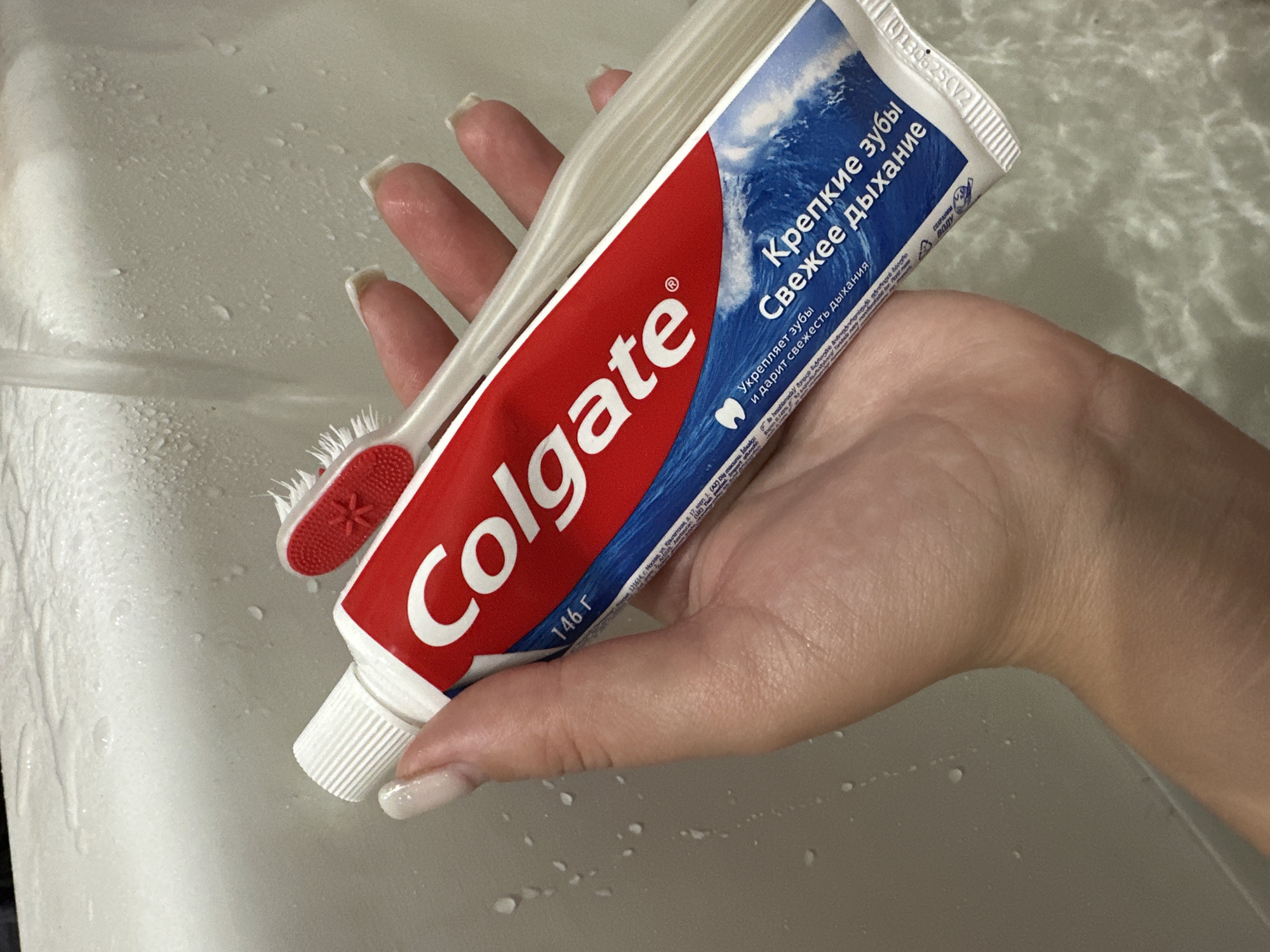 The image shows a close-up view of a person's hand holding a tube of Colgate toothpaste and a toothbrush. The hand has long, light-colored fingernails and is positioned over a light-colored surface, possibly a sink or bathtub, with water droplets visible. The Colgate tube is red and white with blue text and a picture of waves. The toothbrush is white with a red bristle head. The scene appears to be in a bathroom environment, but no specific location in Novokuznetsk, Russia can be determined from the image.