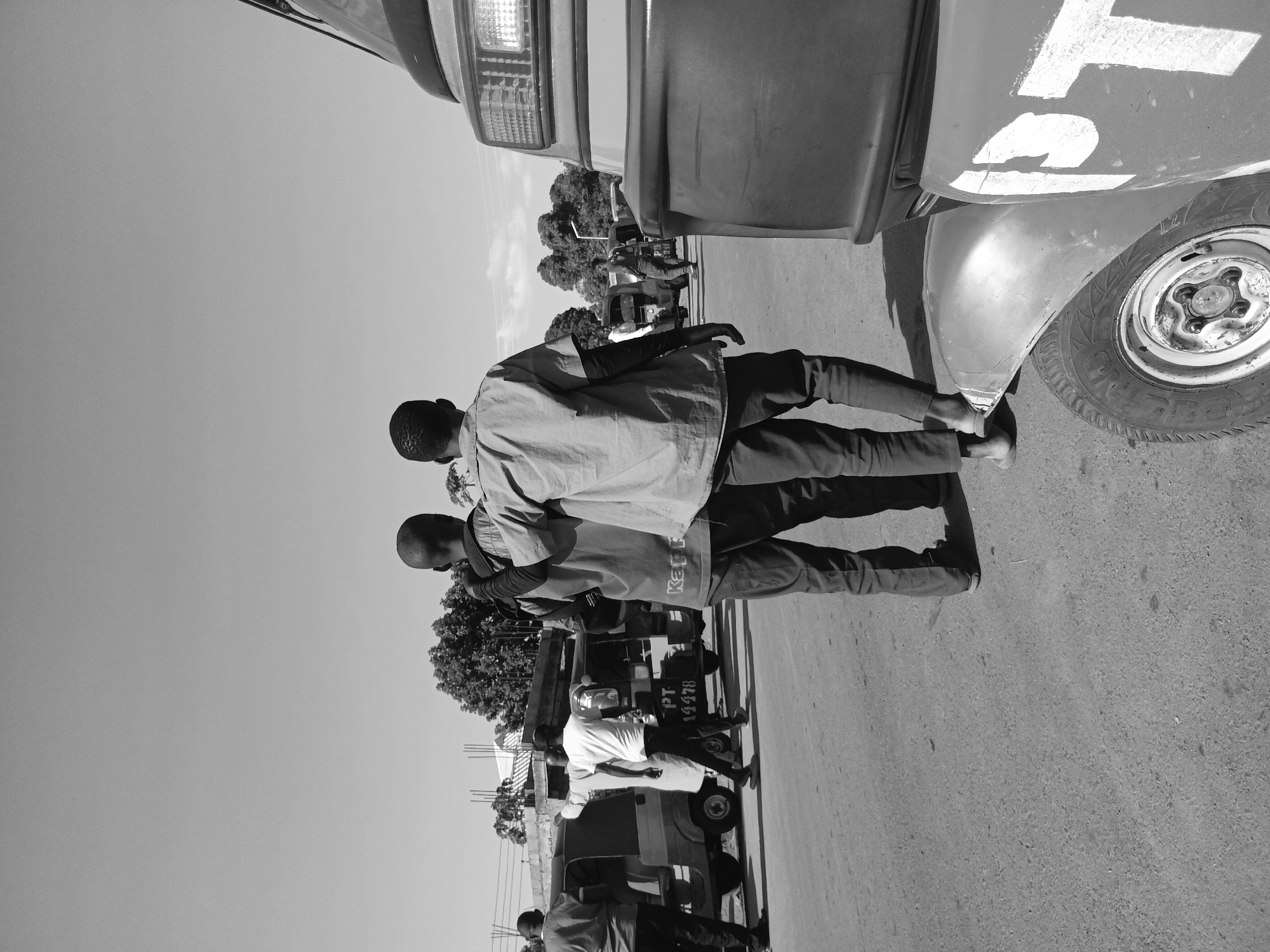 In Nabatum, Nigeria, two young men walk away from the camera, one with his arm around the other's shoulder. They are on a dusty, paved road, with several auto-rickshaws and other vehicles parked or moving in the background. The sky is clear and bright. The image is a black and white photograph, taken from a low angle, with the foreground dominated by the back of an auto-rickshaw. The men appear to be walking towards a busy street, suggesting a commercial or transportation hub. The general atmosphere is one of everyday life in a Nigerian city, with a focus on human interaction and movement.