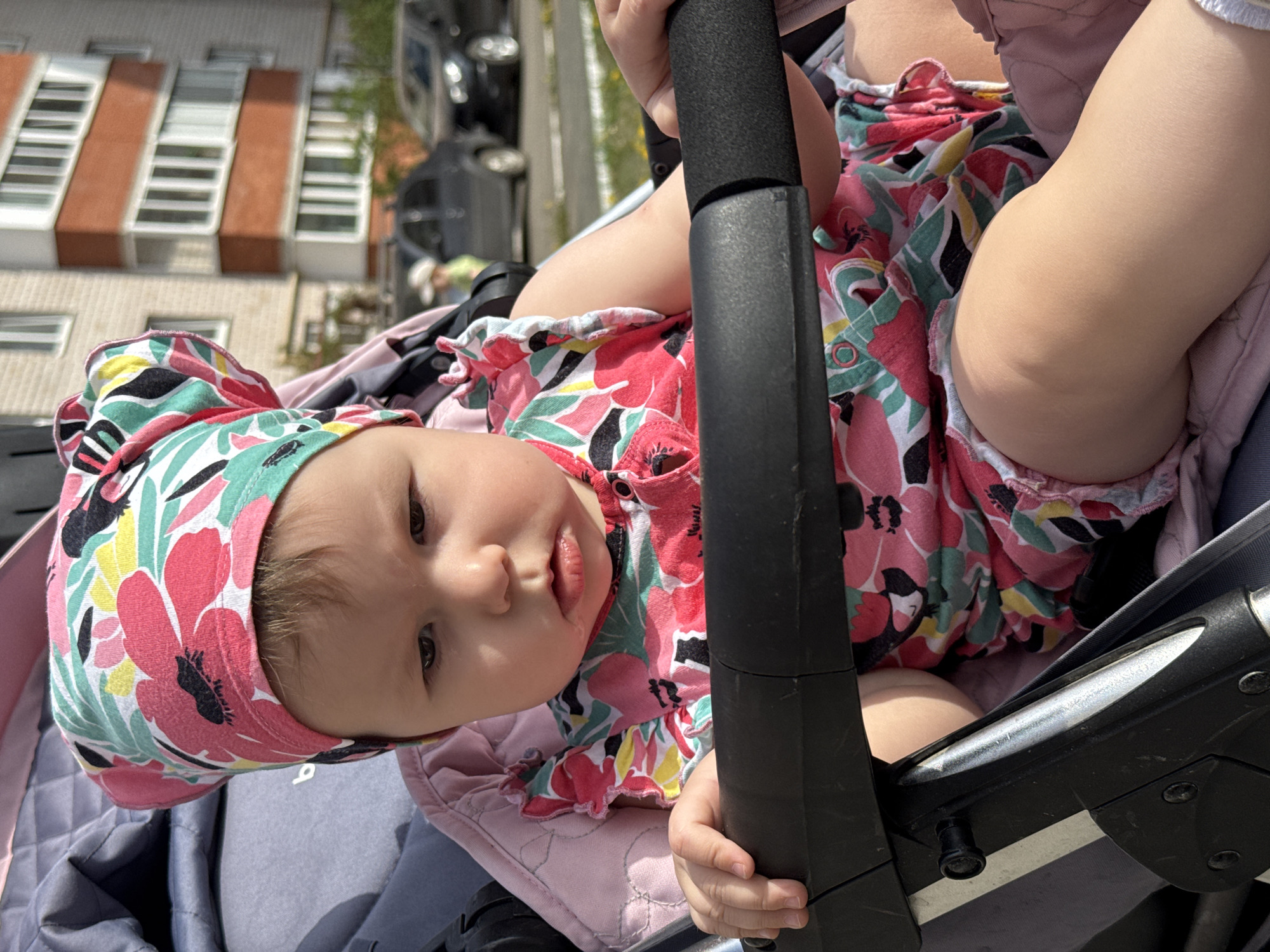 A baby sits in a stroller outdoors, possibly in Zav'yalovo, Russia. The baby wears a floral print hat and matching outfit. The stroller is black, and the baby is holding onto the handle. A building with white and red brick is in the background.
