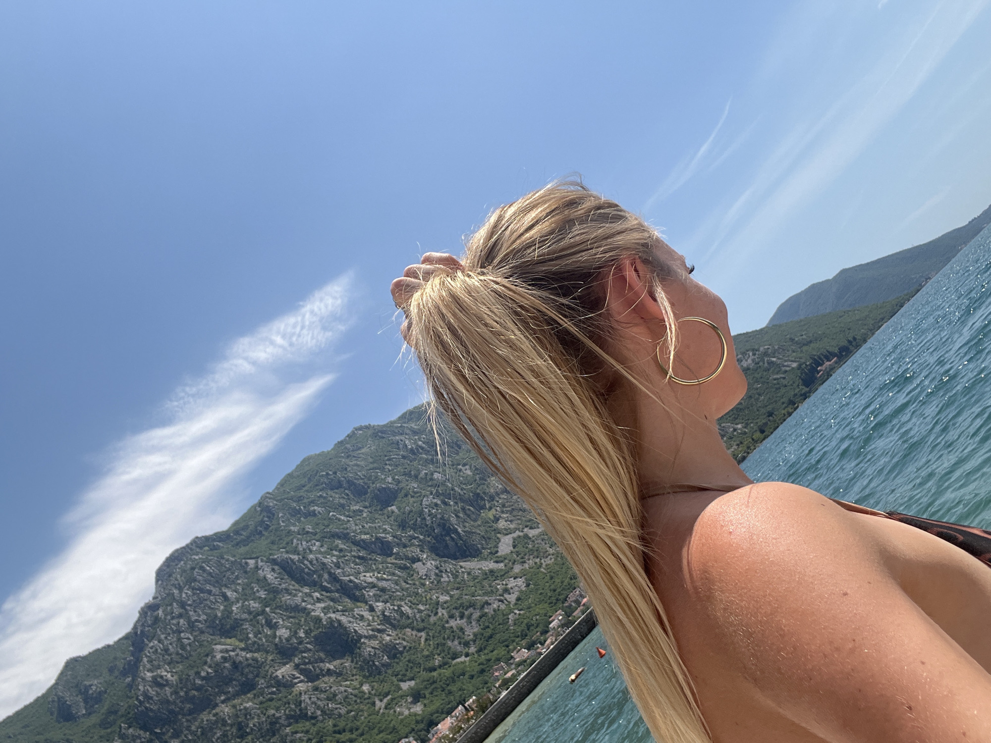 A young woman with blonde hair pulled into a ponytail, wearing a gold hoop earring, looks down and to the right. She has a tan and appears to be on a boat on the water. The background features a steep, green, mountainous coastline with buildings nestled at its base, under a clear blue sky with wispy white clouds. This scene is consistent with the scenery found in Risan, Montenegro, known for its dramatic bay and coastal mountains.