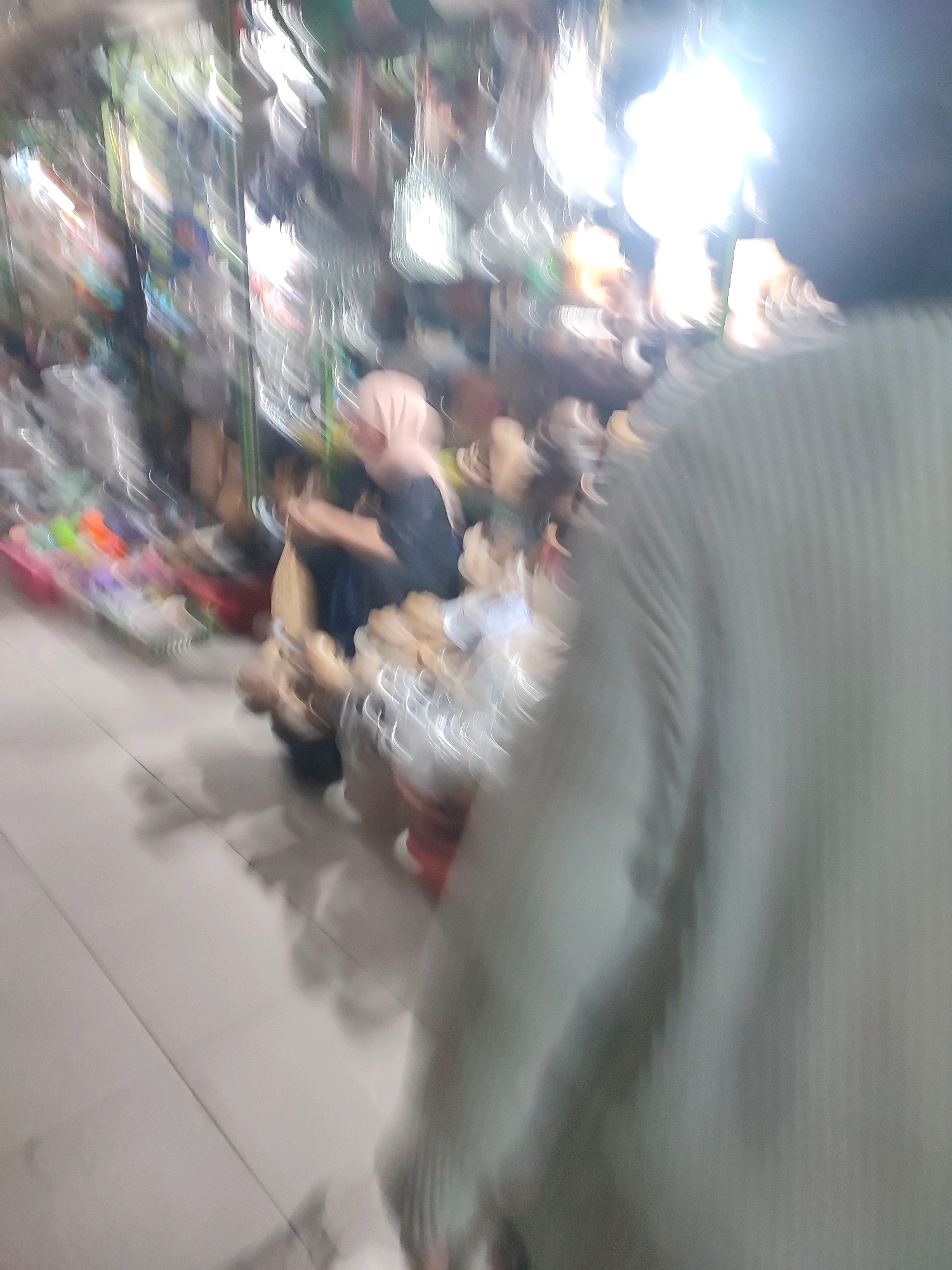 The image depicts a bustling street market scene in Yogyakarta, Indonesia. The foreground is dominated by the back of a person wearing a light-colored, ribbed sweater, suggesting they are moving away from the camera. The background is a blur of activity, with stalls and people visible. One stall appears to be selling various items, possibly textiles or small crafts, with a rack of colorful objects to the left. A woman wearing a headscarf and a dark top is visible behind a counter, interacting with customers. Another stall features what look like shoes or sandals, displayed on shelves. The lighting is bright, with a strong glare in the upper right portion of the image, indicating overhead lighting or sunlight. The overall impression is one of a lively and crowded marketplace, characteristic of many urban centers in Indonesia.