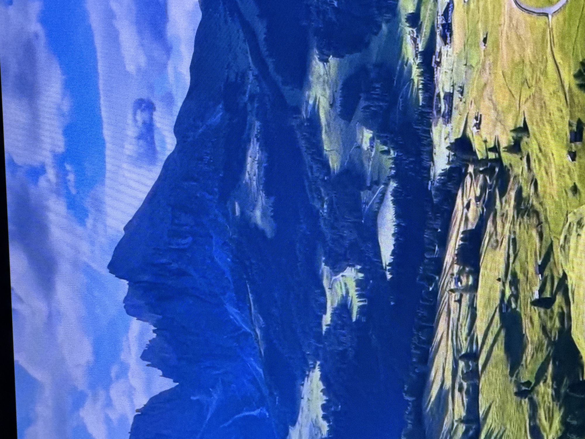 The image displays a striking optical illusion where a mountain range is shaped to resemble a human profile, specifically a face in silhouette. The "face" is dark blue, with distinct features such as a prominent nose, lips, and chin, against a backdrop of soft blue and white clouds. The mountain's surface suggests rugged, textured terrain, with some areas appearing lighter due to shadows or sunlight.
To the right of the mountainous profile, the scene transitions to a vibrant green landscape, indicative of rolling hills or meadows. This area is marked with darker patches, likely trees or shadowed areas, creating a textured visual. A paved, curved pathway is visible in the upper right portion of this green area, suggesting a man-made element within the natural scenery. The lighting suggests it is daytime, with shadows indicating the sun's position.
The image is a photograph, possibly taken on a screen or display, as indicated by the slightly pixelated appearance and the texture of the surface. The orientation is such that the mountain profile is on the left and the green landscape is on the right, with the image rotated such that the "face" appears to be looking left. There is no visible text in the image. The overall impression is one of natural beauty and artistic illusion, blending geological formations with a human likeness.