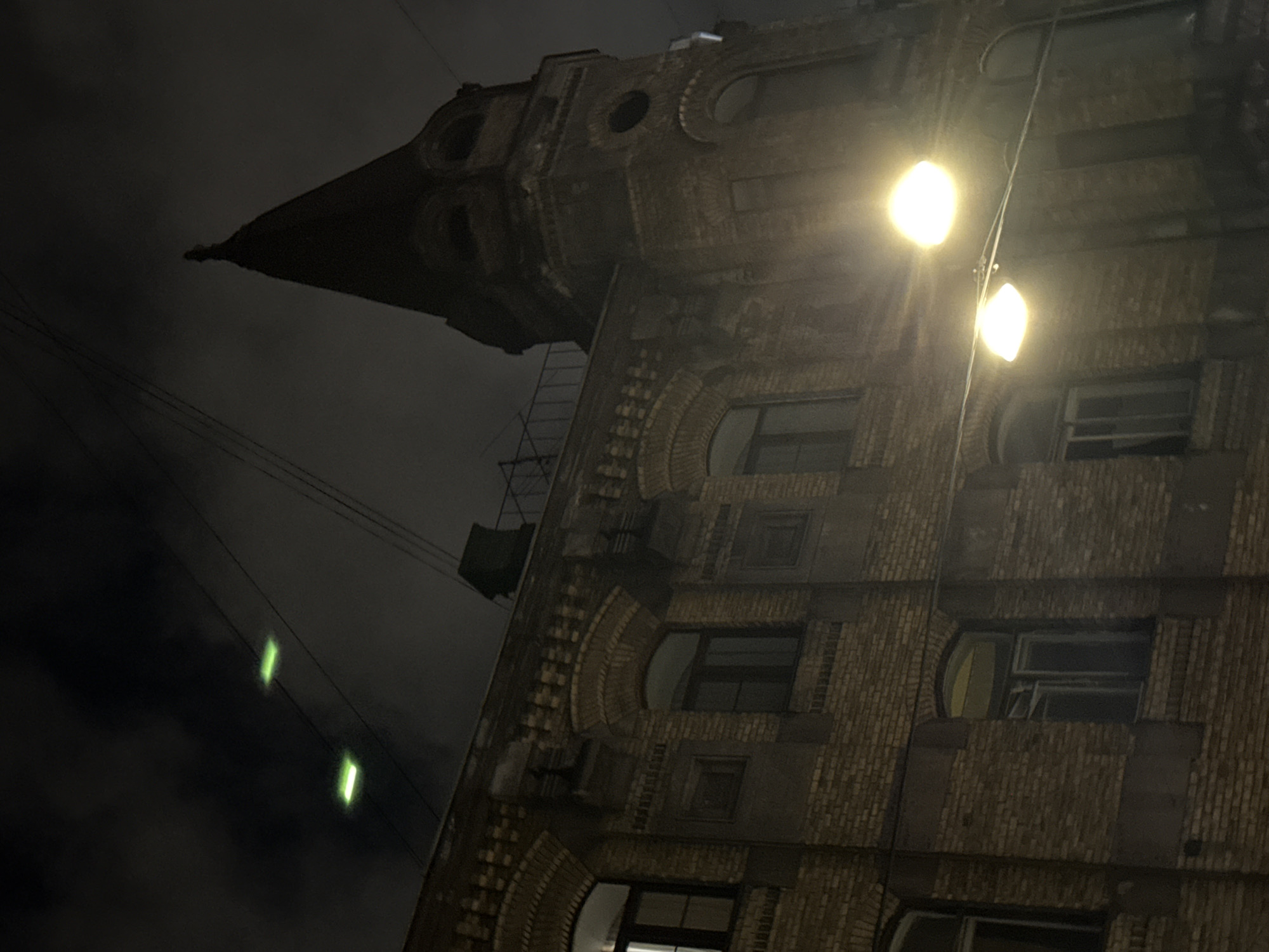Low-angle night shot of a tall brick building in Saint Petersburg, Russia. The building has a distinctive triangular peak and rounded windows. A ladder and other infrastructure are visible on the side of the building. Two streetlights illuminate the building's facade. Green lights are faintly visible in the distance, possibly from a transmission line. The sky is dark and cloudy.