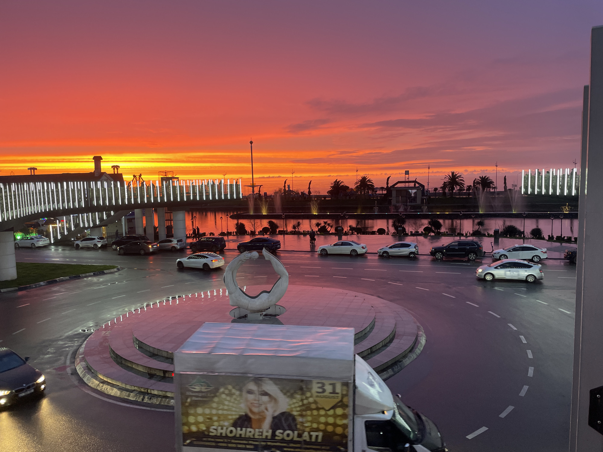 The image displays an urban twilight scene in Batumi, Georgia, overlooking a section of the city's modern boulevard area near the Black Sea. The sky is dominated by intense hues of orange and red at the horizon, transitioning to purple and dark blue overhead, indicating either a strong sunset or sunrise.
In the foreground, a multi-lane asphalt road, visibly wet and reflecting ambient light, forms a large roundabout. A prominent, abstract sculpture, white in color and resembling two interlocking rings, is centrally located within the roundabout. Multiple vehicles are present on the wet pavement: a dark-colored car with headlights illuminated is visible in the lower-left corner, and various sedans and SUVs, predominantly white or dark-colored (including what appears to be a white Tesla Model 3 and a silver hatchback), are circulating or positioned on the lanes. A white delivery truck with a large advertisement for "SHOHREH SOLATI" printed on its side is partially visible in the lower-right foreground.
Beyond the road and roundabout, a body of water features multiple illuminated fountains. A modern pedestrian bridge, characterized by numerous vertical white LED light panels, spans across the scene. Palm trees and other urban structures line the waterfront promenade. In the background, silhouetted against the vivid sky, are the outlines of a Ferris wheel and other amusement park structures, typical of the Batumi Boulevard. No individuals are clearly discernible in the scene.