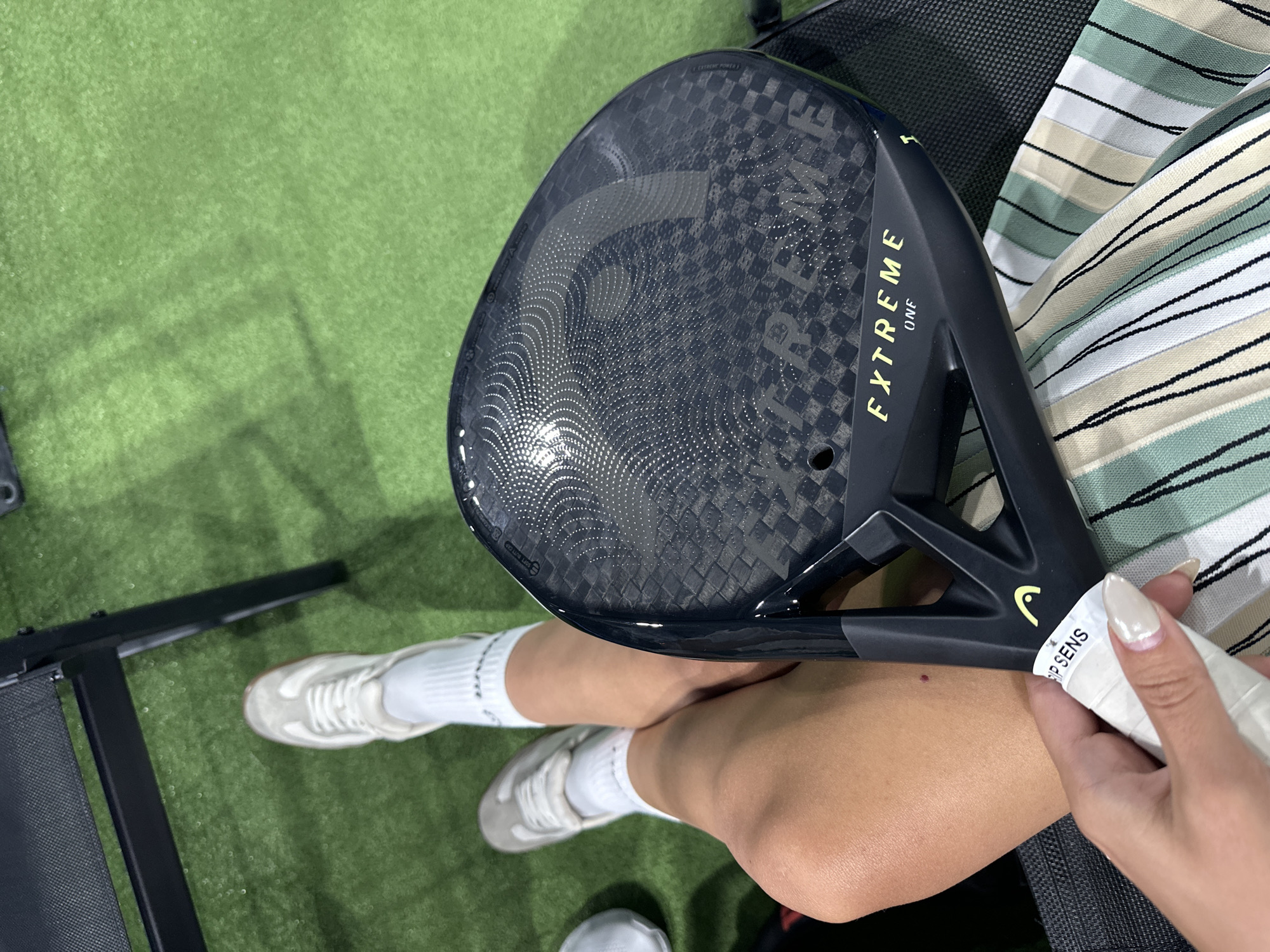 A person is sitting on a green artificial turf surface, holding a black padel racket. The racket has "EXTREME ONE" written in yellow on it. The person is wearing white socks and light-colored sneakers. Their legs are crossed. A black chair is partially visible in the background. The scene is indoors, possibly at a sports facility. There is no indication of the city of Sofia, Bulgaria.