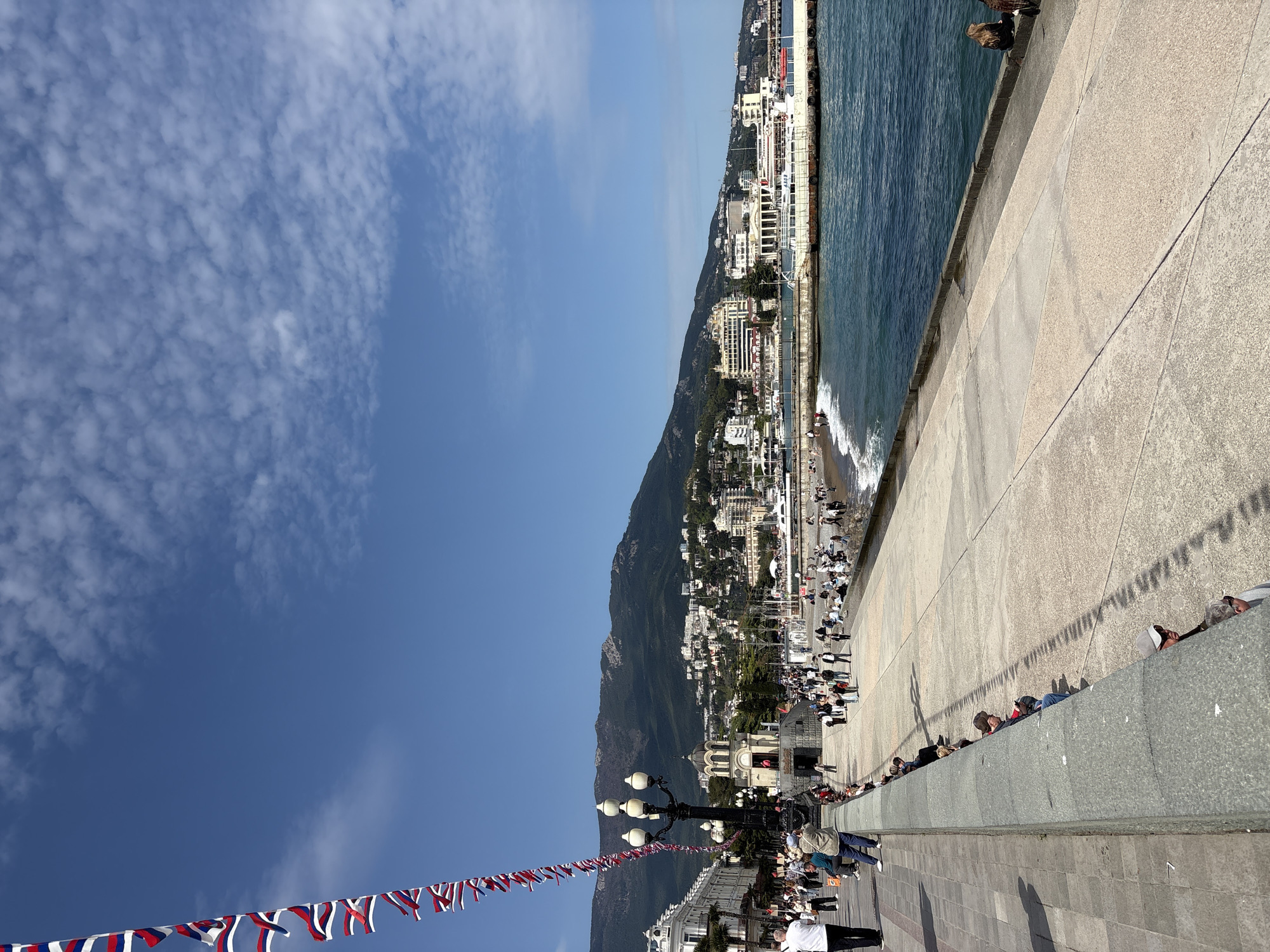 The image depicts a coastal scene in Ялта, Ukraine. A concrete embankment runs along the edge of the sea. People are seen walking and sitting along the embankment's edge, some looking out to the water. A mountainous area rises behind the city, which consists of various buildings. A string of flags, displaying the colors of the Russian flag, adorn the scene. The sky is blue with scattered clouds. The perspective suggests the photo was taken from an elevated position looking downwards.