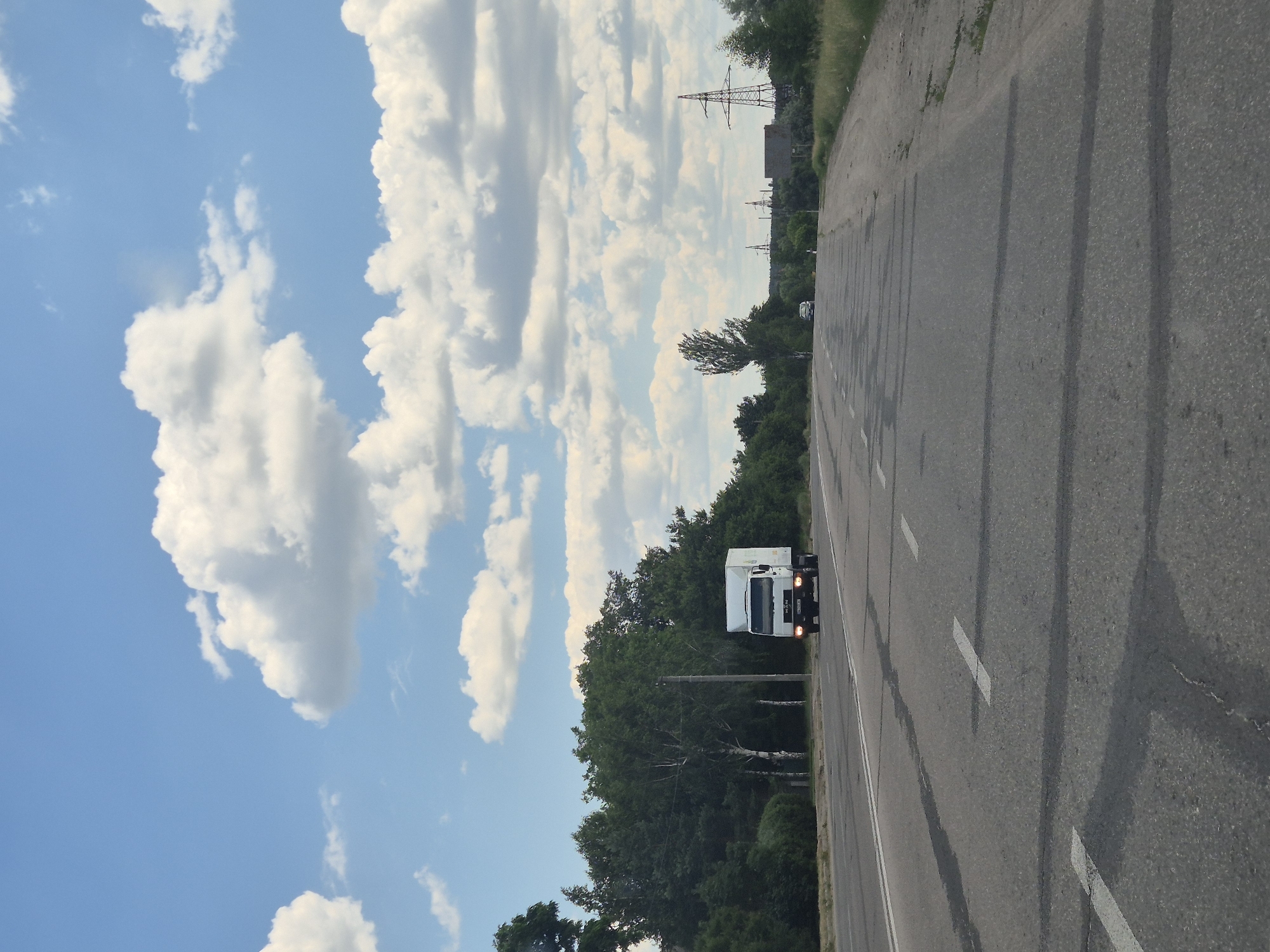 A white truck with a box trailer is driving on a paved road in Pidhaitsi, Ukraine. The road has white lane markings and appears to be a two-lane highway. To the left of the road is a dense line of green trees, including some birch trees. To the right of the road, there is a grassy shoulder and more trees in the distance. In the far background, there are power lines and what appear to be industrial structures. The sky is blue with scattered white, fluffy clouds. The lighting suggests it is daytime, likely late afternoon due to the warm tone of the light.
