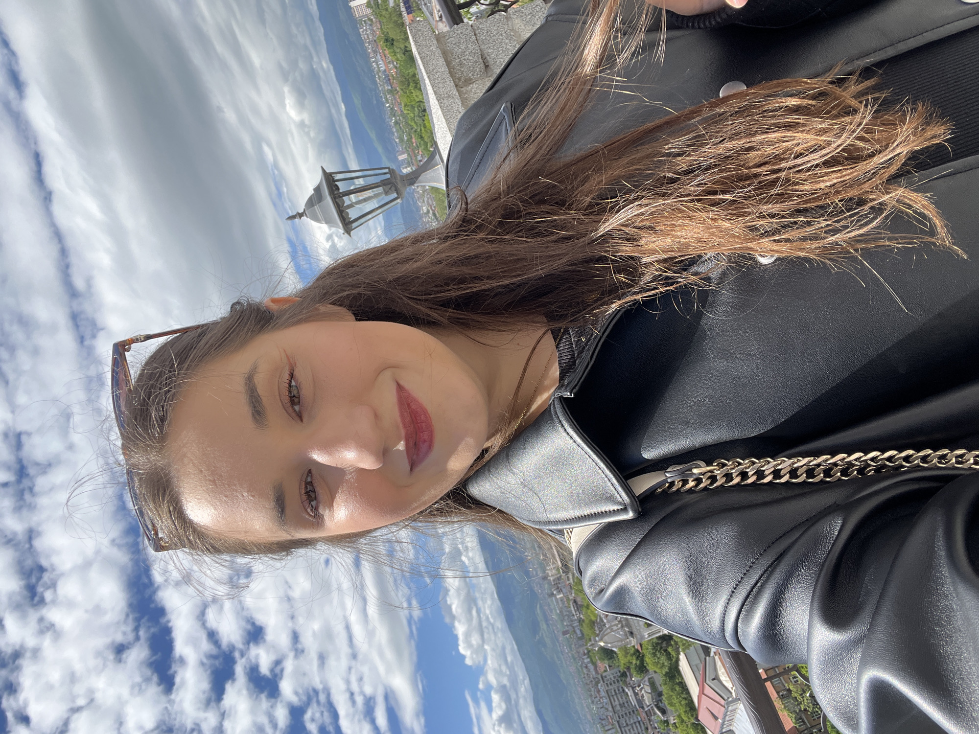 A young woman is taking a selfie while overlooking the city of Plovdiv, Bulgaria. The photo is taken from a high vantage point, showing a panoramic view of the city. The woman is wearing a black leather jacket and has brown hair. In the background, there are buildings, mountains, and a cloudy blue sky. A lamppost is also visible. The woman's face is partially visible. She has sunglasses propped on her head. Her hair is long, straight, and brown, and she is wearing lipstick. The image is taken during daylight hours.